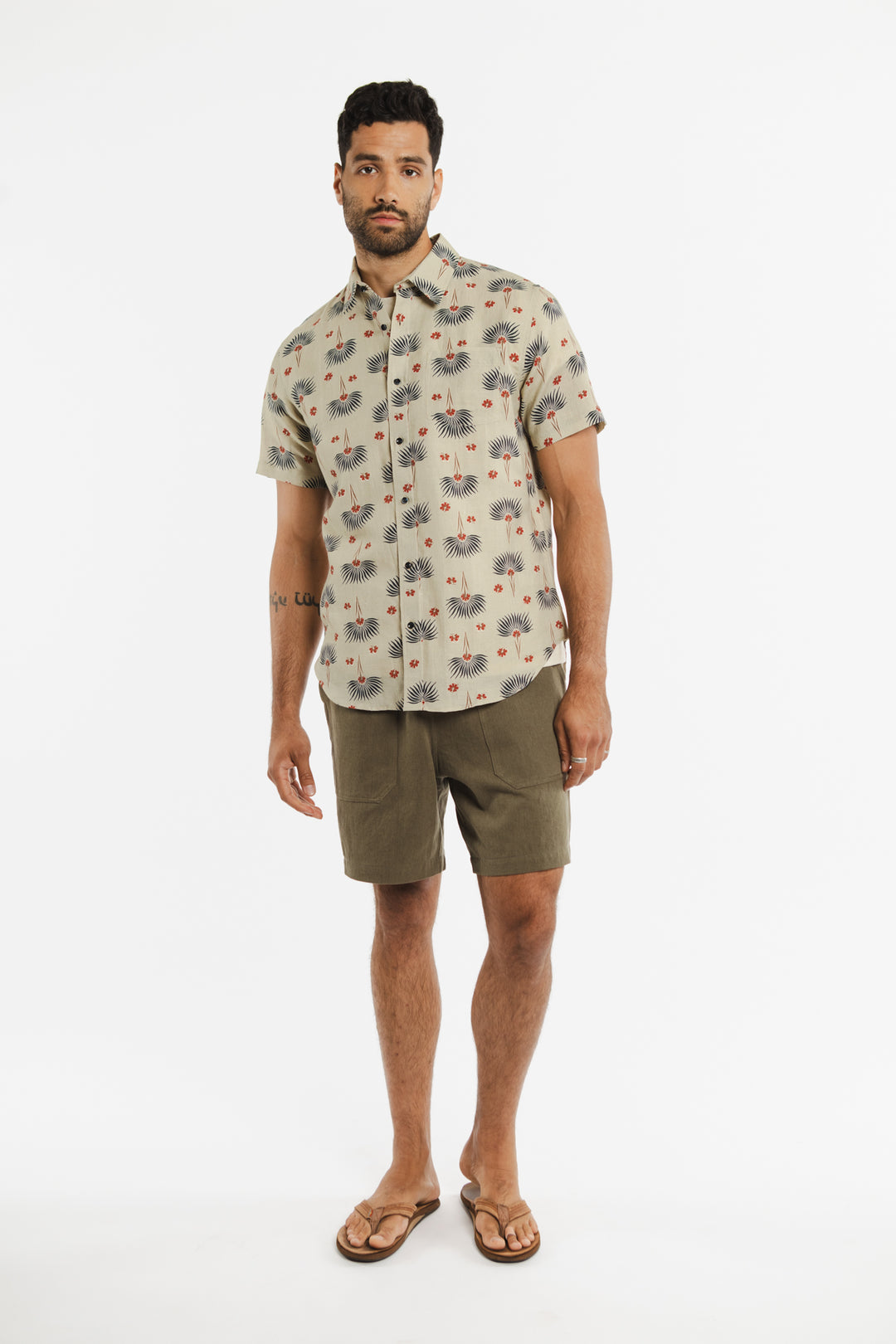 A man with a short beard and curly hair stands against a white background, wearing the Bridge & Burn Harbor Slim Shirt in Aloha Floral. The shirt is slim fit with short sleeves, paired with olive green shorts and brown flip-flops.