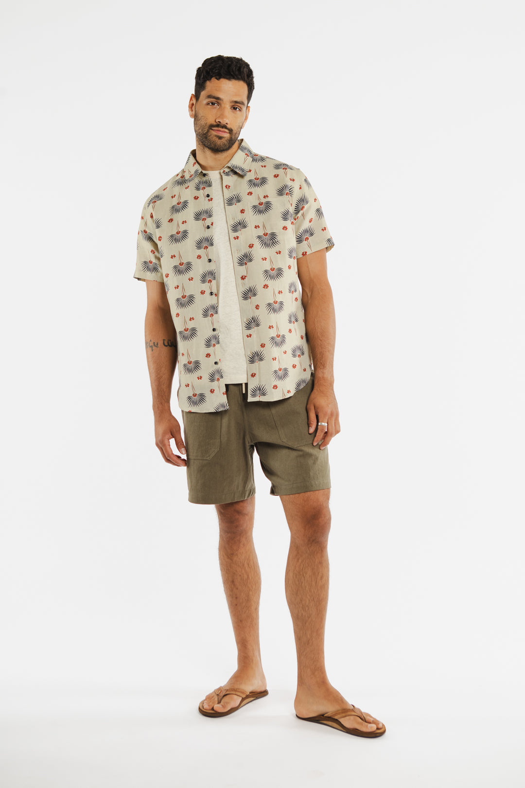 A man with short hair and a beard stands against a white background wearing the Bridge & Burn Harbor Slim Shirt in Aloha Floral, a light t-shirt, olive green shorts, and brown flip-flops with a relaxed posture.