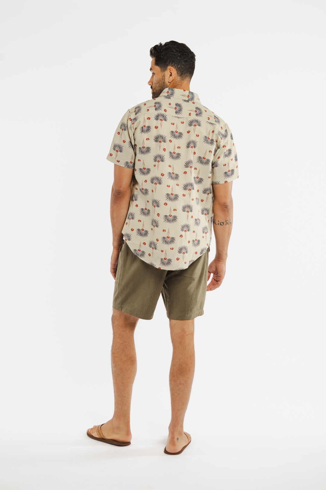 A man stands facing away, wearing the Bridge & Burn Harbor Slim Shirt in Aloha Floral and olive-green shorts with brown sandals, against a plain white background.