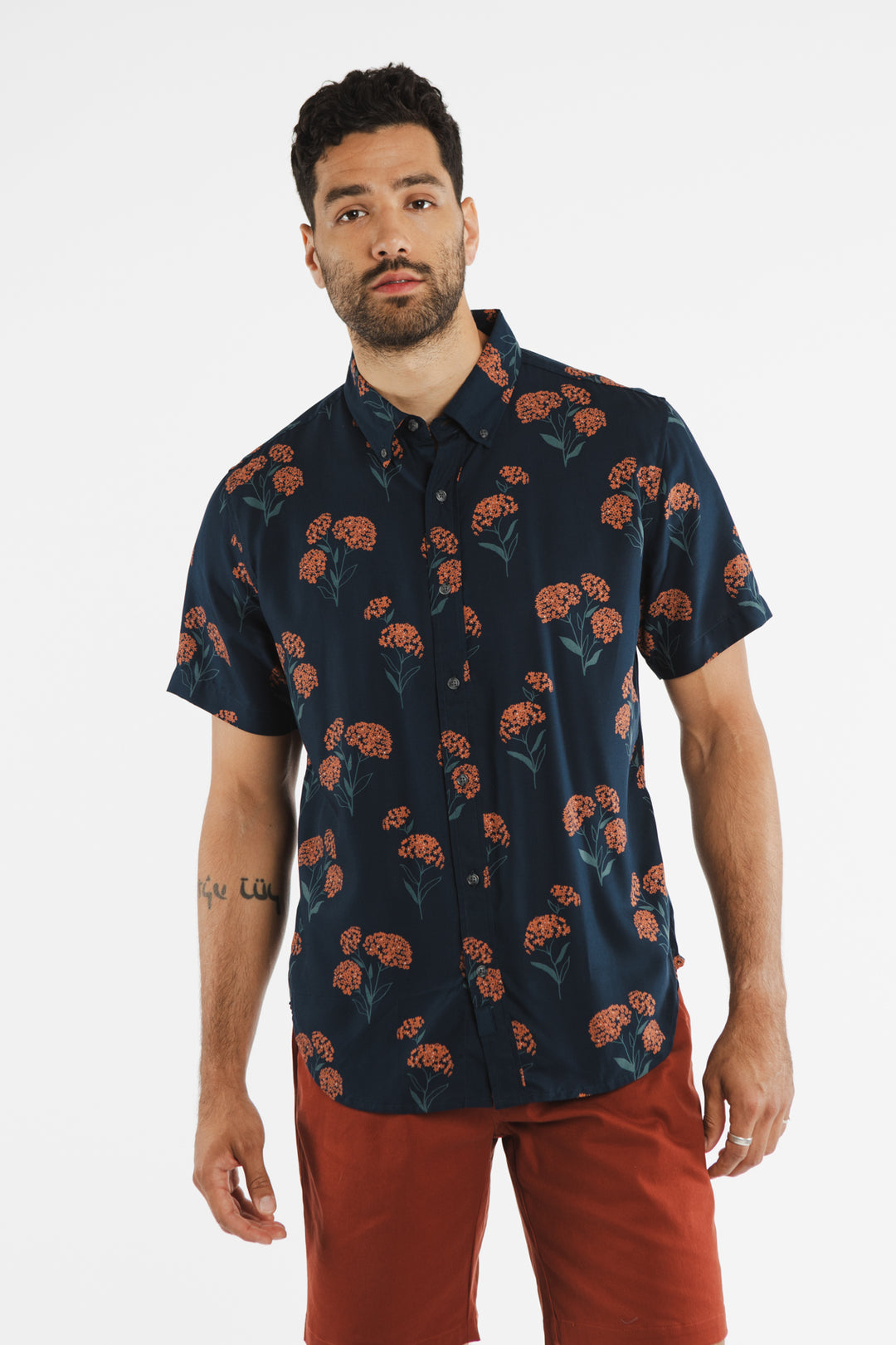 A man with short curly hair and a beard wears the Bridge & Burn Grant Slim Shirt / Wildflower with an orange floral pattern and rust-colored shorts, standing against a plain white background.