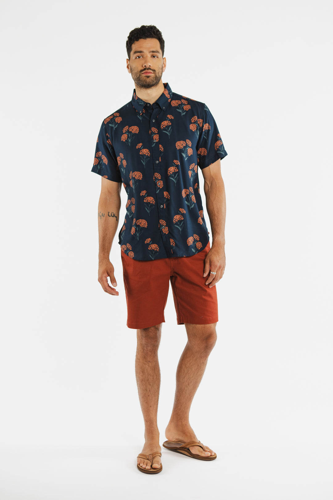A man with short hair and a beard stands against a white background wearing the Bridge & Burn Grant Slim Shirt in Wildflower, paired with rust shorts and tan flip-flops. One arm is slightly bent.