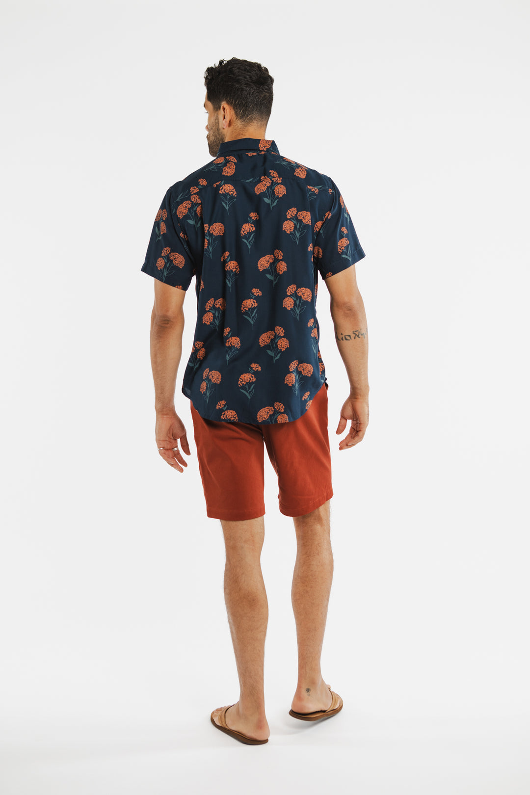 A man faces away in a Bridge & Burn Grant Slim Shirt / Wildflower—a navy slim-fit shirt with an orange floral pattern—paired with burnt orange shorts and flip-flops, set against a simple white background.