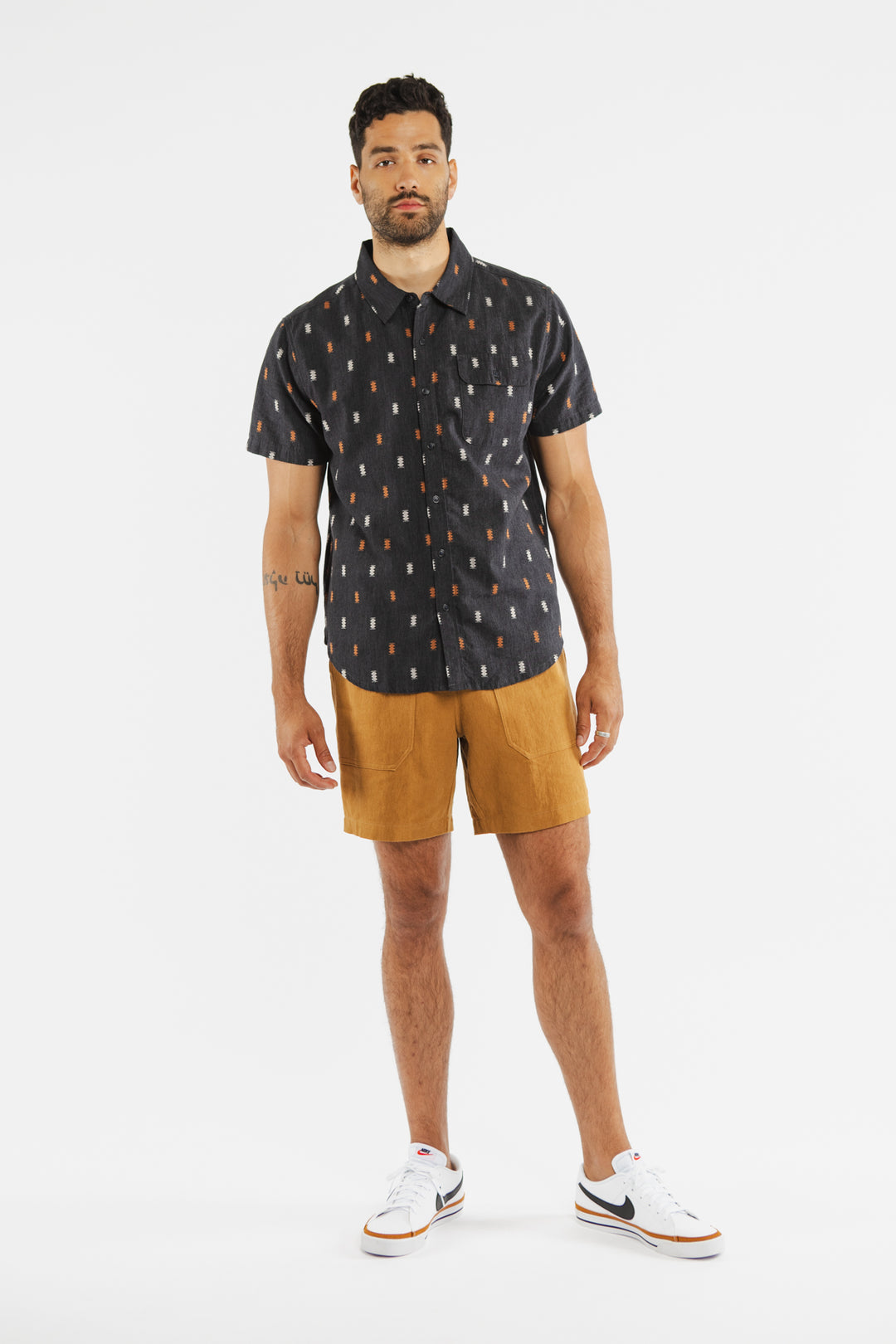 A man with short hair and a trimmed beard stands against a white background wearing Bridge & Burn's Noah Short in Ochre, a black patterned short-sleeve button-up shirt, and white sneakers with orange accents.