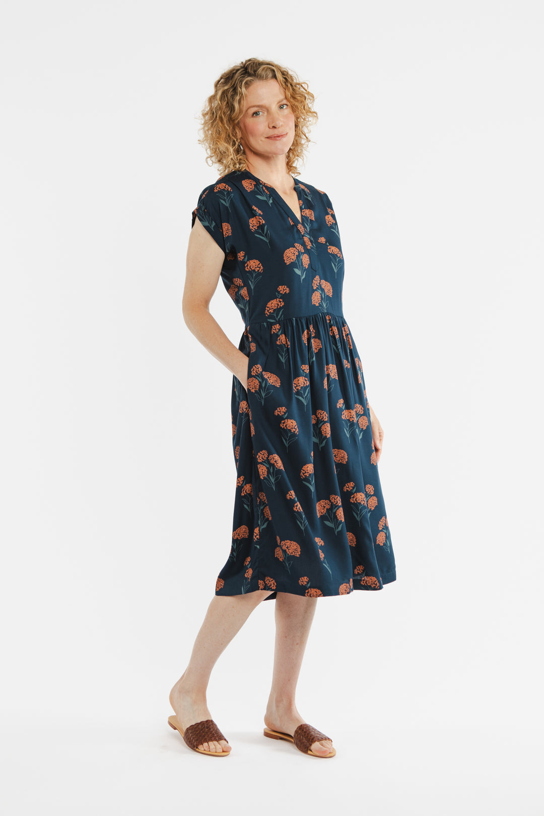 A person with curly hair is wearing the Bridge & Burn Ira Dress in Wildflower, a patterned, navy blue, knee-length pullover with short sleeves and pockets. They stand against a plain white background, one hand in a pocket.
