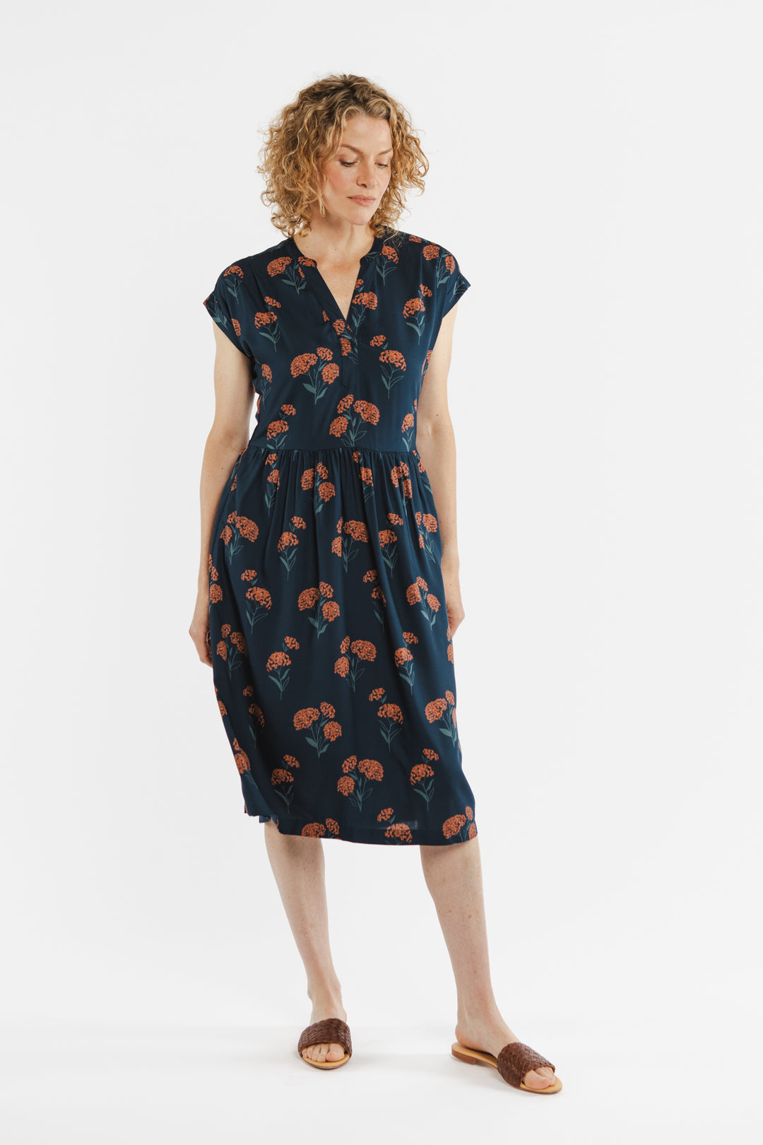 A woman with curly hair models the Bridge & Burn Ira Dress in Wildflower—a navy knee-length pullover with a red floral print. She stands against a white background, looking down, hands by her sides, and wears brown sandals.