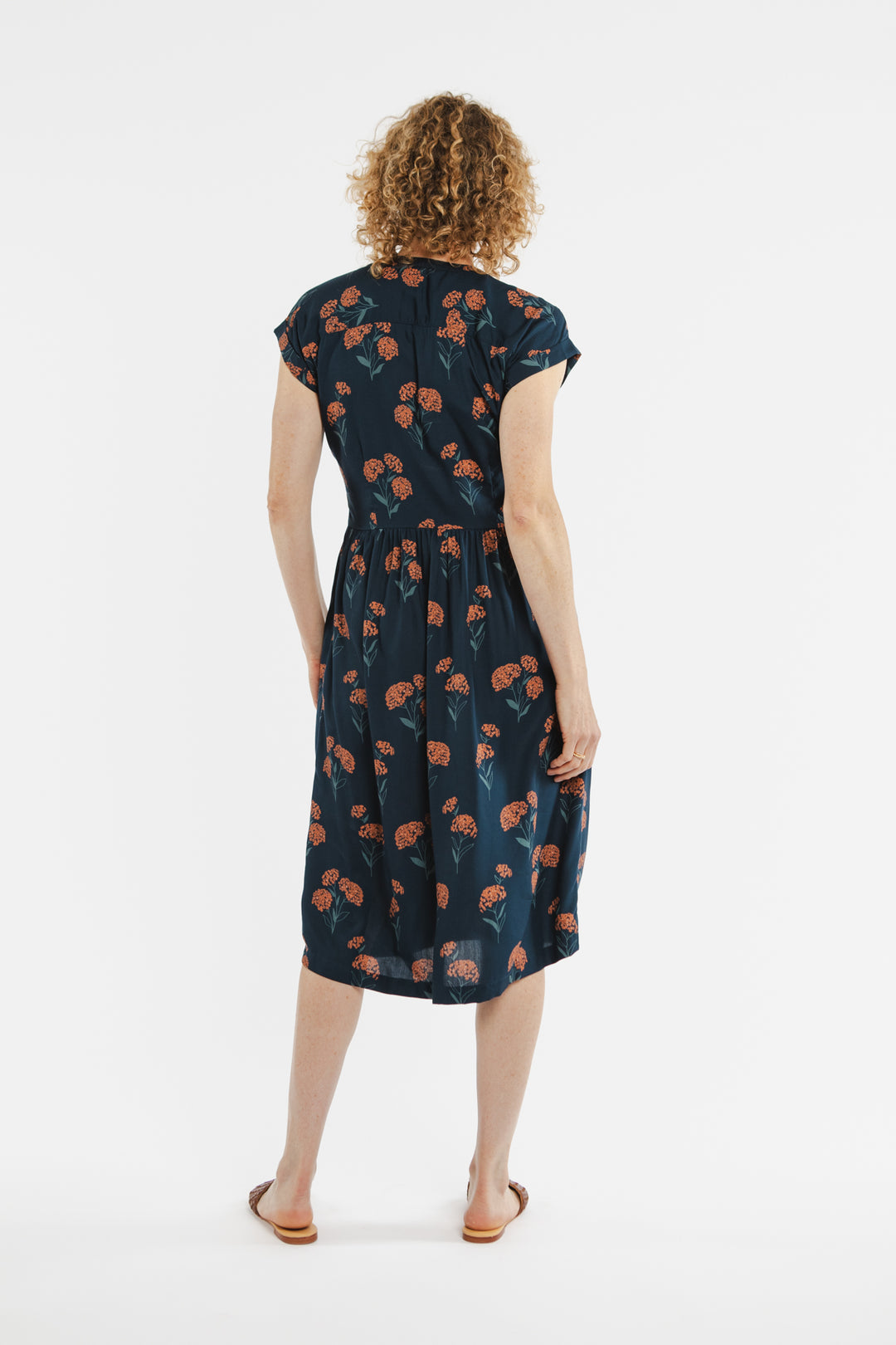A woman with curly hair stands facing away from the camera, wearing the Bridge & Burn Ira Dress in the Wildflower print, styled with flat sandals, against a plain white background.