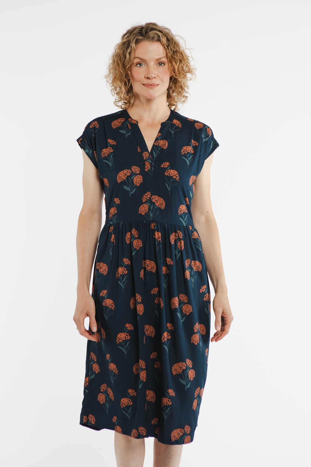 A woman with curly blonde hair stands against a white background, wearing the Bridge & Burn Ira Dress in the Wildflower print.