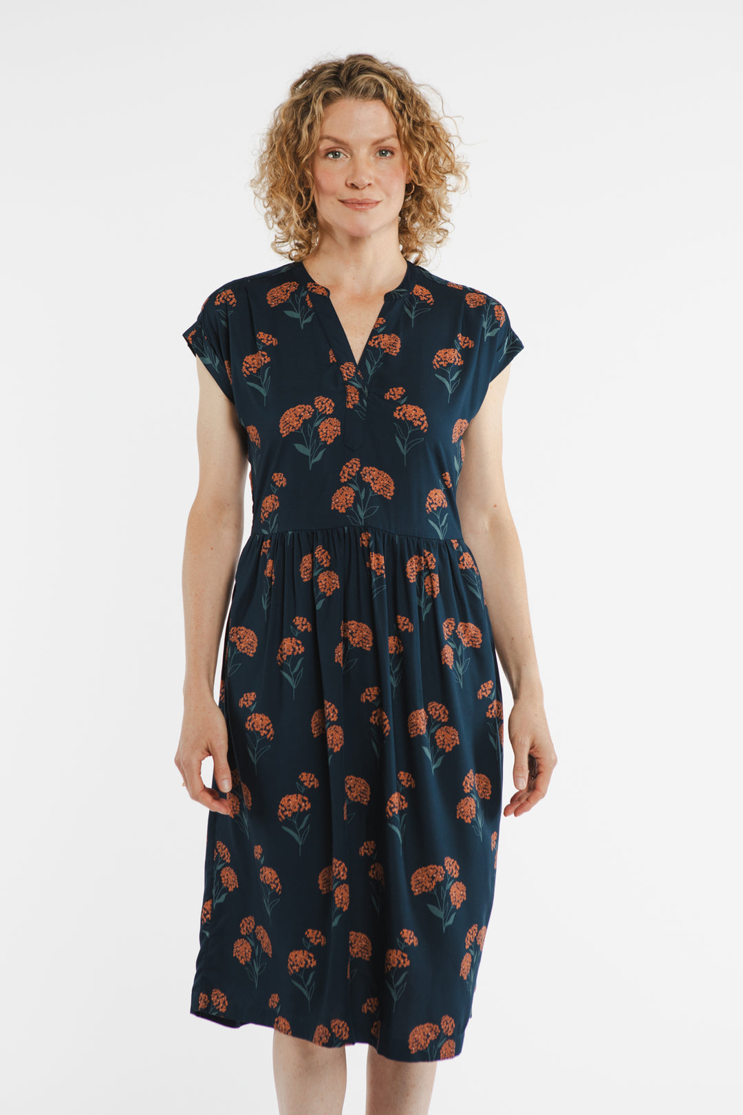A woman with curly blonde hair stands against a white background, wearing the Bridge & Burn Ira Dress in the Wildflower print.