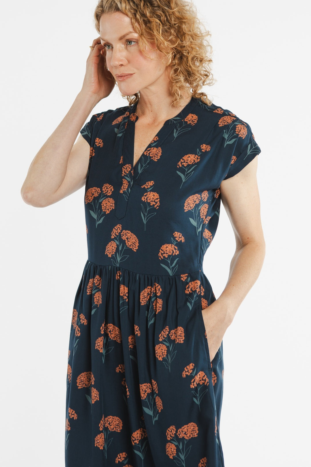 A woman with curly blonde hair wears the Bridge & Burn Ira Dress in Wildflower, a navy blue modal dress with an orange floral pattern. She stands against a plain white background, touching her hair and looking to the side.