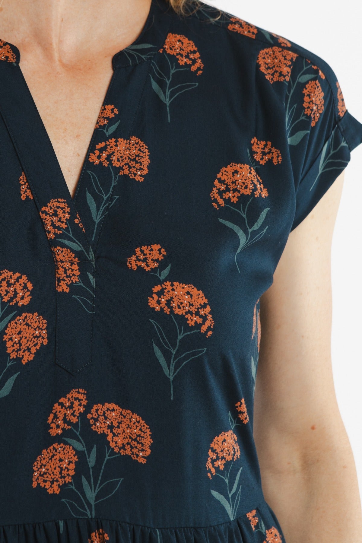 A close-up of a person wearing the Bridge & Burn Ira Dress in Wildflower, a navy blue pullover dress with a v-neckline, short sleeves, and an orange floral cluster pattern with green stems and leaves.