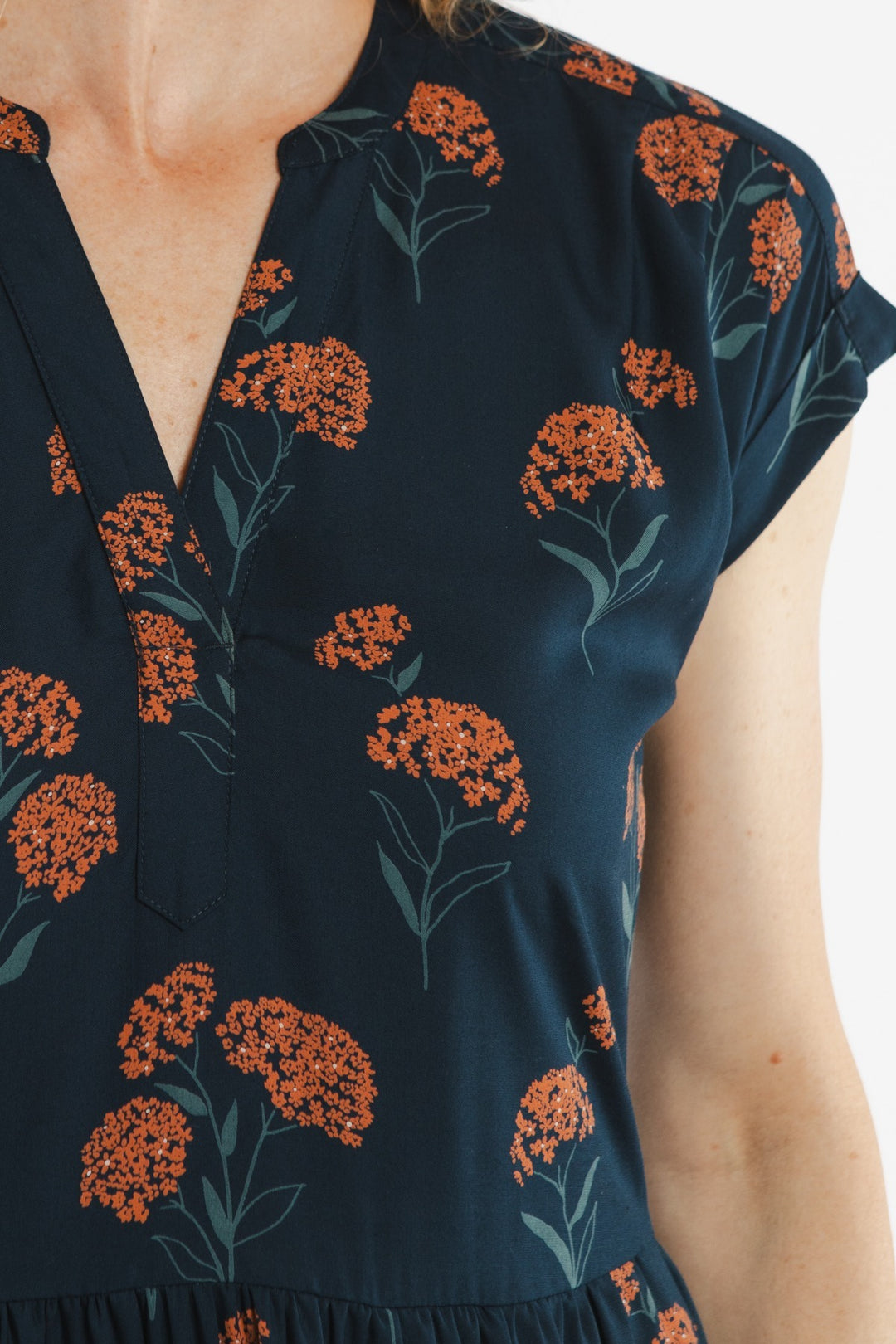 A close-up of a person wearing the Bridge & Burn Ira Dress in Wildflower, a navy blue pullover dress with a v-neckline, short sleeves, and an orange floral cluster pattern with green stems and leaves.