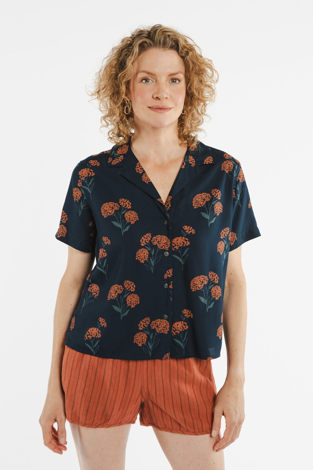 A person with curly blonde hair wears the Bridge & Burn Mila Cropped Shirt in the Wildflower pattern and rust-colored shorts, standing against a plain white background.