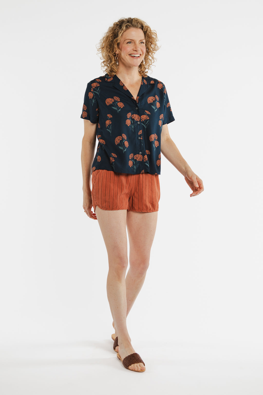 A smiling person with curly hair wears the Bridge & Burn Mila Cropped Shirt in Wildflower, paired with rust shorts and brown sandals, walking against a plain white background.