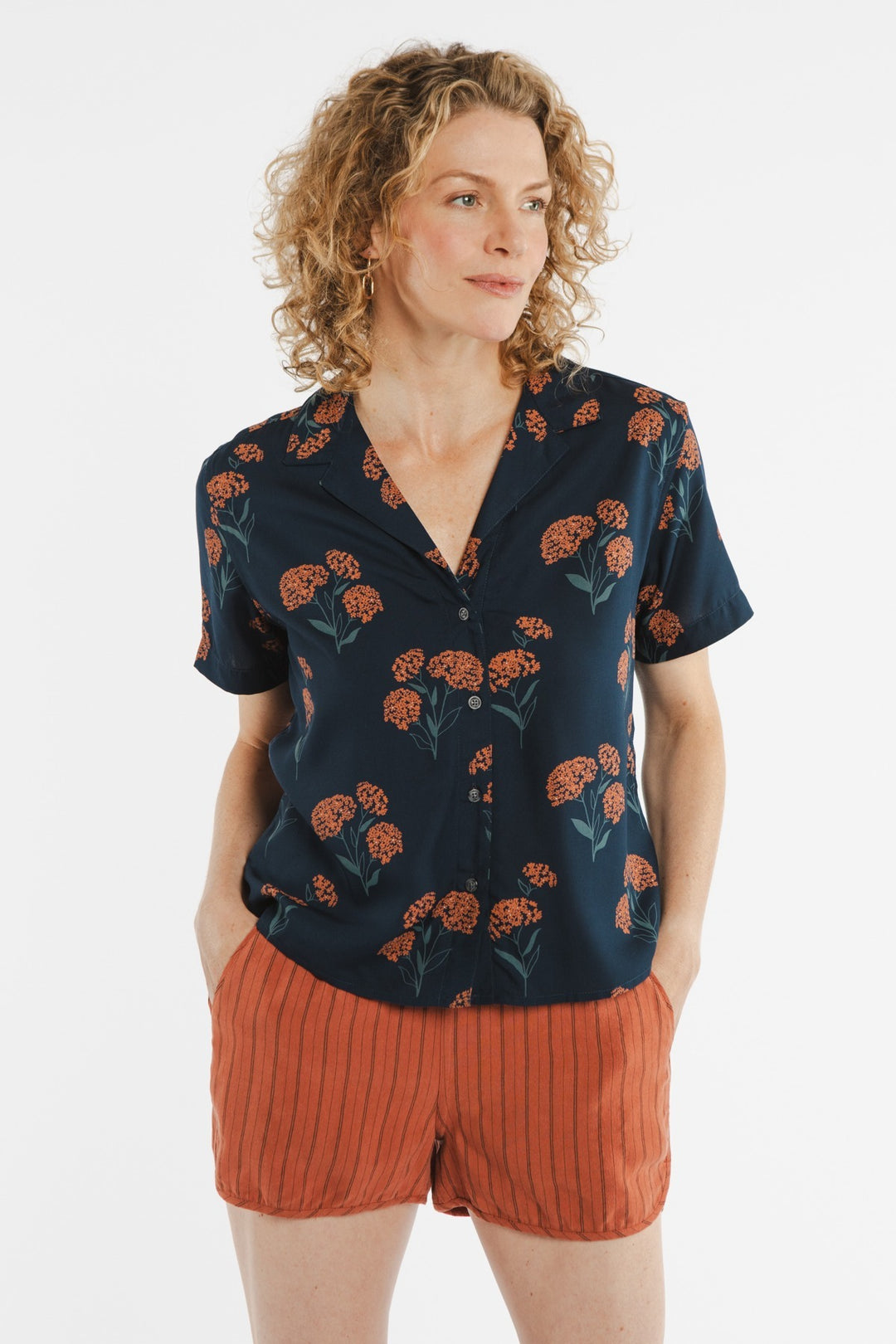 A woman with curly blond hair wears the Bridge & Burn Mila Cropped Shirt in Wildflower, styled with rust-colored striped shorts and her hands in her pockets, standing against a white background.