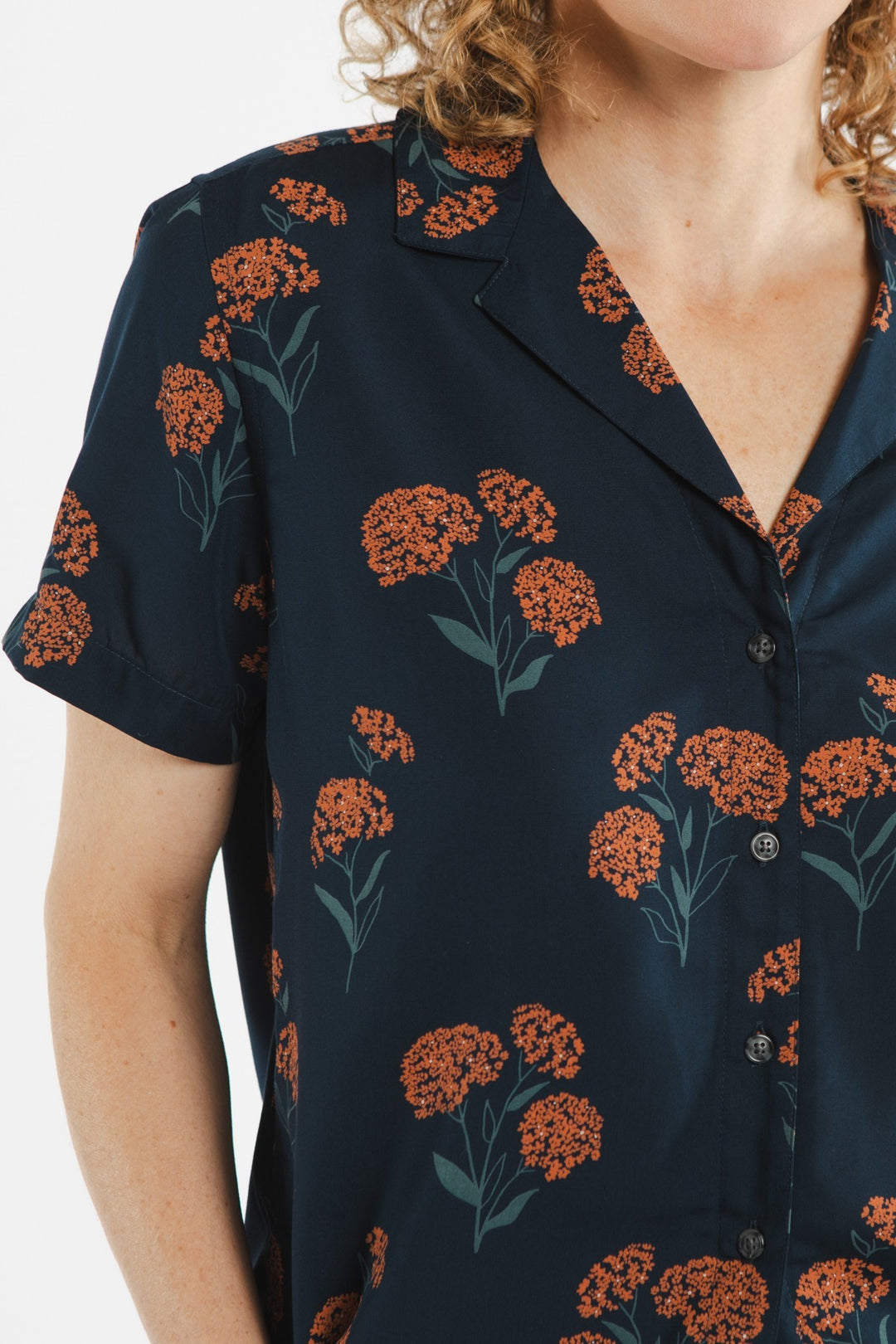 A person wears the Bridge & Burn Mila Cropped Shirt in Wildflower, a dark navy modal top with an orange floral print, collar, front buttons, and short sleeves; only their upper torso and part of their face are visible.