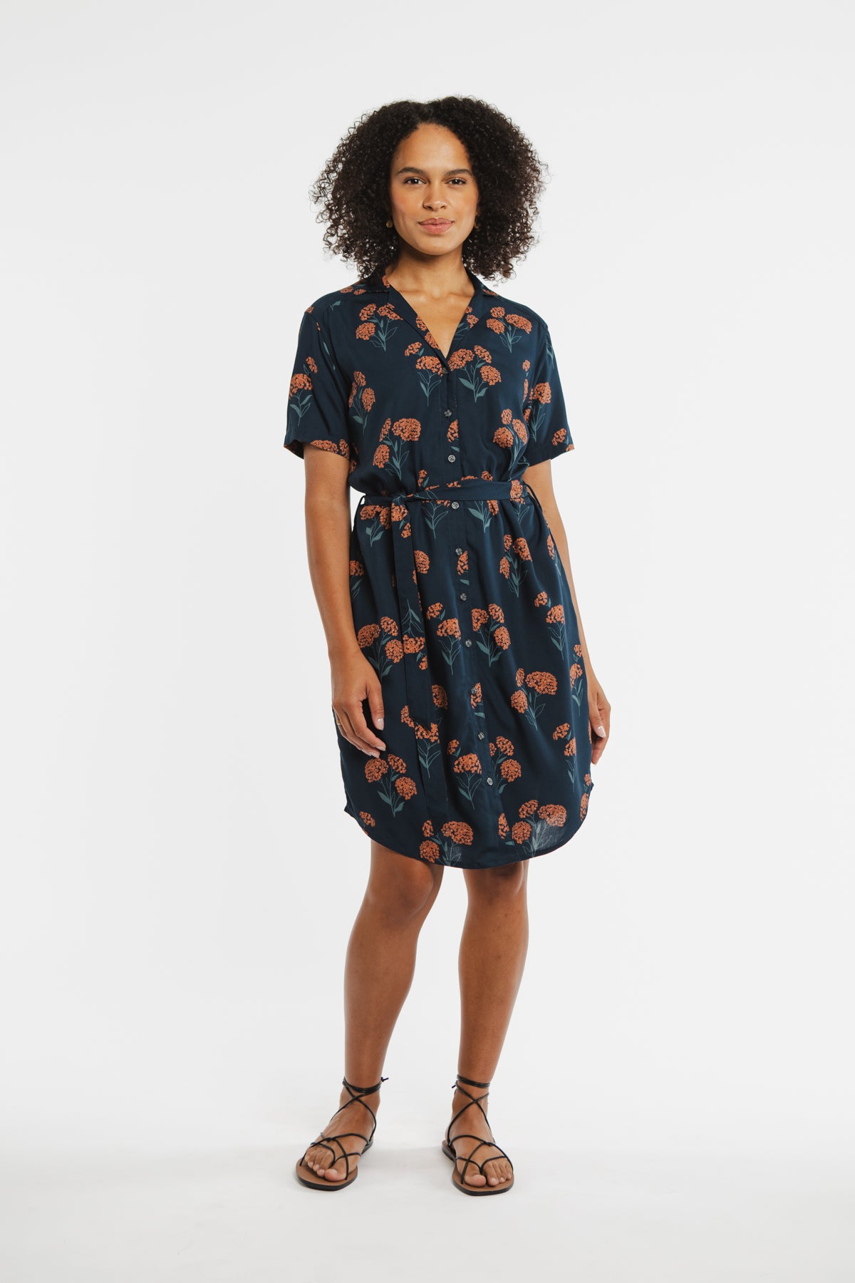 A person stands against a white background wearing the Bridge & Burn Sofia Shirt Dress in Wildflower—a navy blue, self-belted button-front dress with an orange floral print—paired with black strappy sandals.
