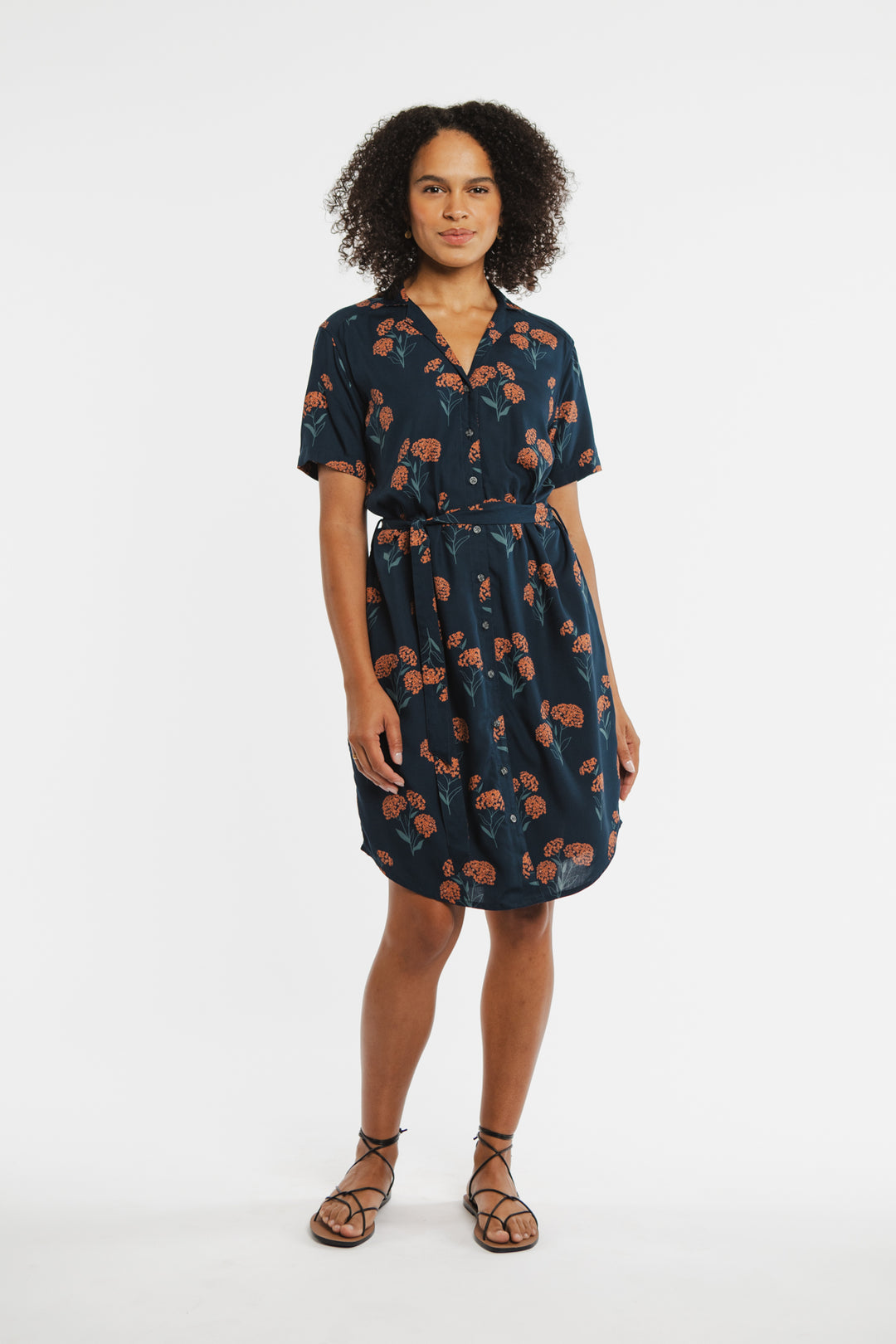 A person stands against a white background wearing the Bridge & Burn Sofia Shirt Dress in Wildflower—a navy blue, self-belted button-front dress with an orange floral print—paired with black strappy sandals.
