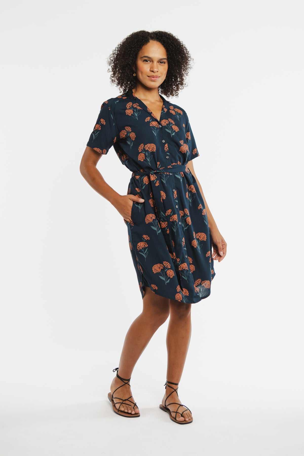 A woman stands confidently in the Bridge & Burn Sofia Shirt Dress / Wildflower, a navy blue self-belted button-front dress with an orange floral print. She has curly hair, wears flat strappy sandals, and poses against a plain white background.