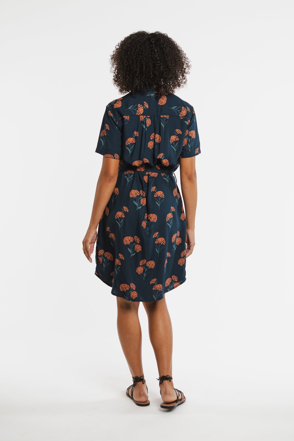 A woman with curly hair faces away, wearing the Bridge & Burn Sofia Shirt Dress in Wildflower—a navy sustainable summer dress with an orange floral print—paired with black sandals against a plain white background.