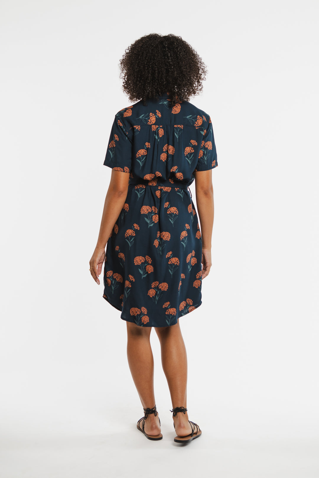 A woman with curly hair faces away, wearing the Bridge & Burn Sofia Shirt Dress in Wildflower—a navy sustainable summer dress with an orange floral print—paired with black sandals against a plain white background.