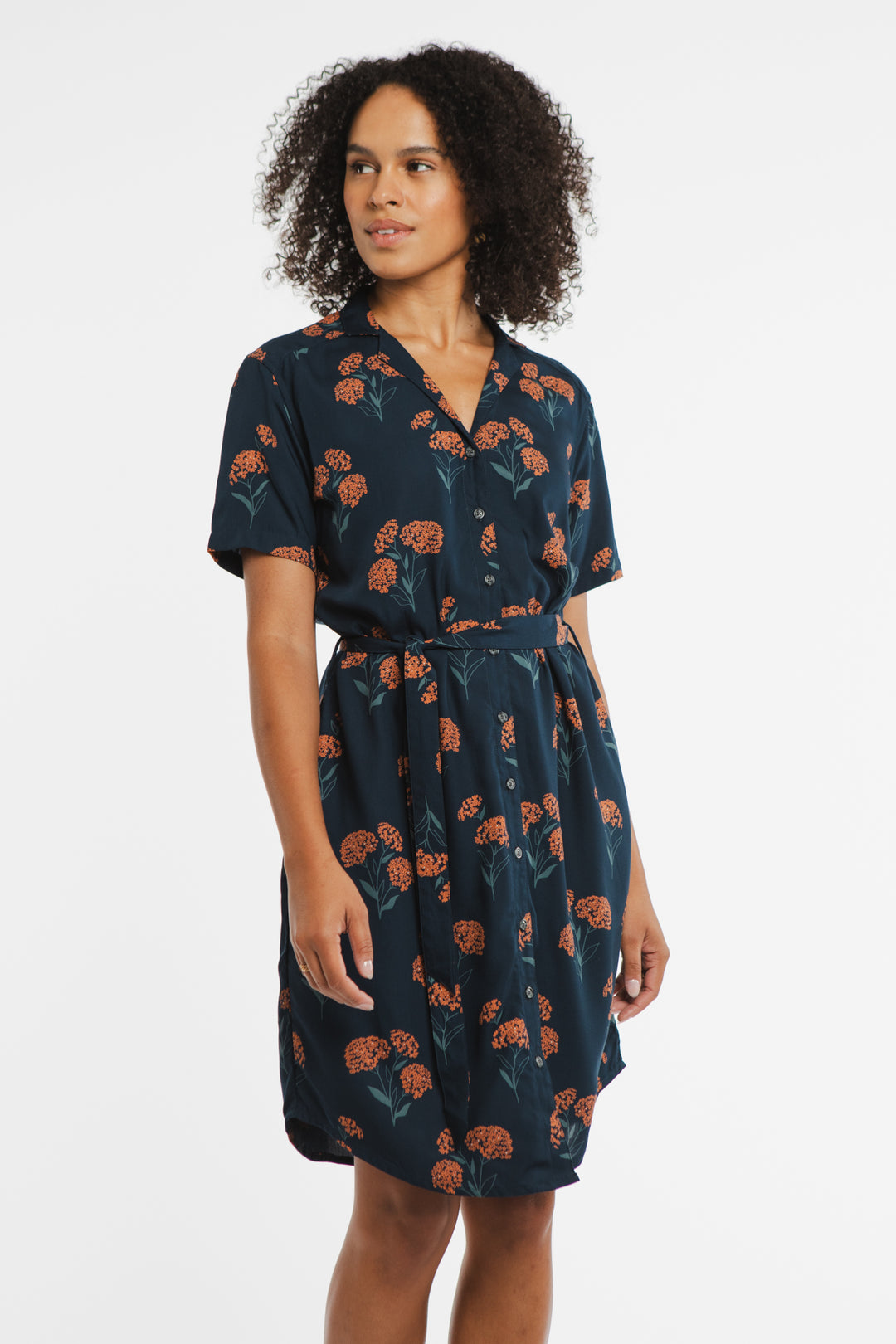 A woman with curly hair wears the Bridge & Burn Sofia Shirt Dress in Wildflower, a navy blue, short-sleeved sustainable summer dress with an orange floral pattern, standing against a plain white background.