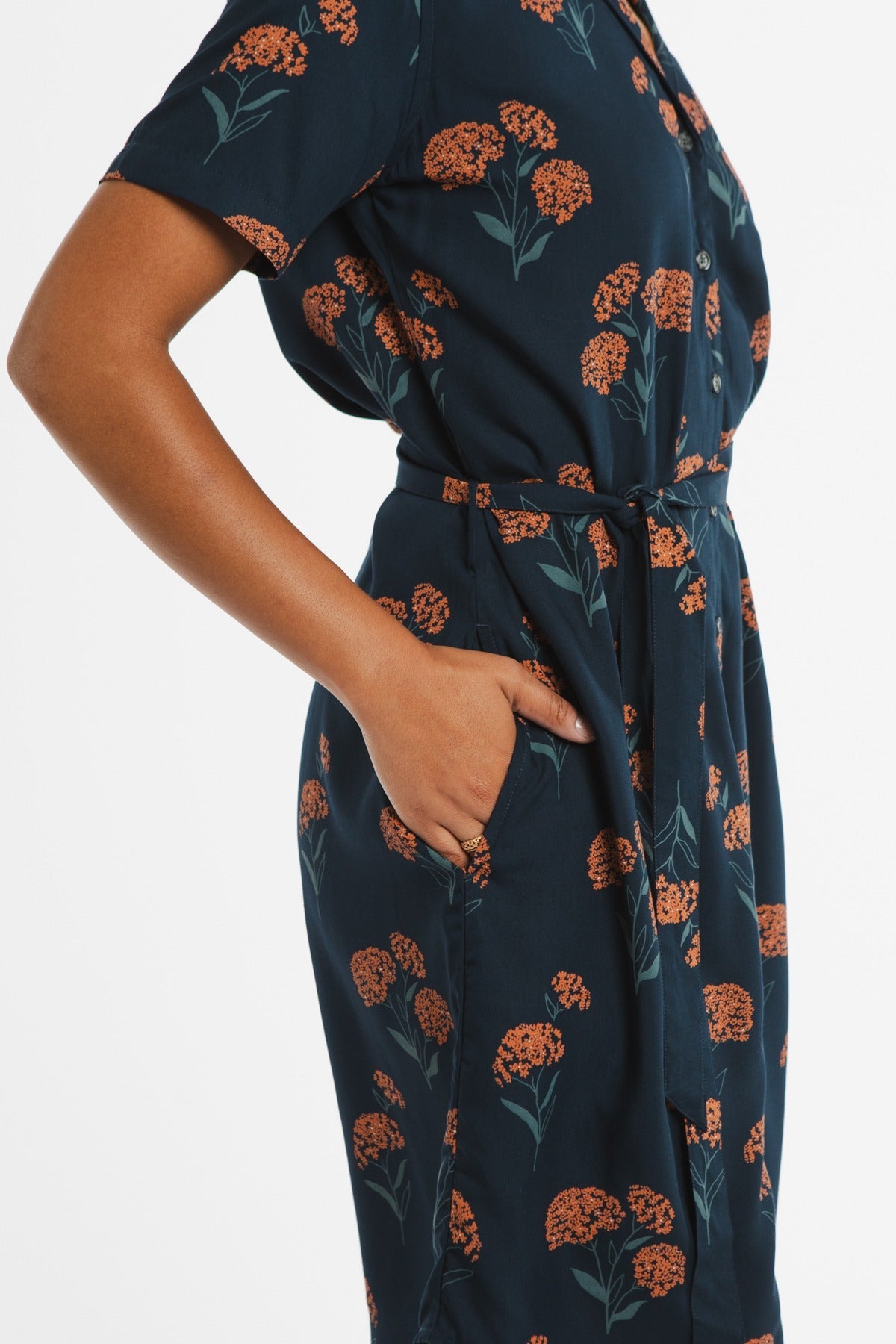 A person wears the Bridge & Burn Sofia Shirt Dress in Wildflower, featuring orange floral patterns, short sleeves, buttons, a self-belt, and side pocket. The background is plain white.
