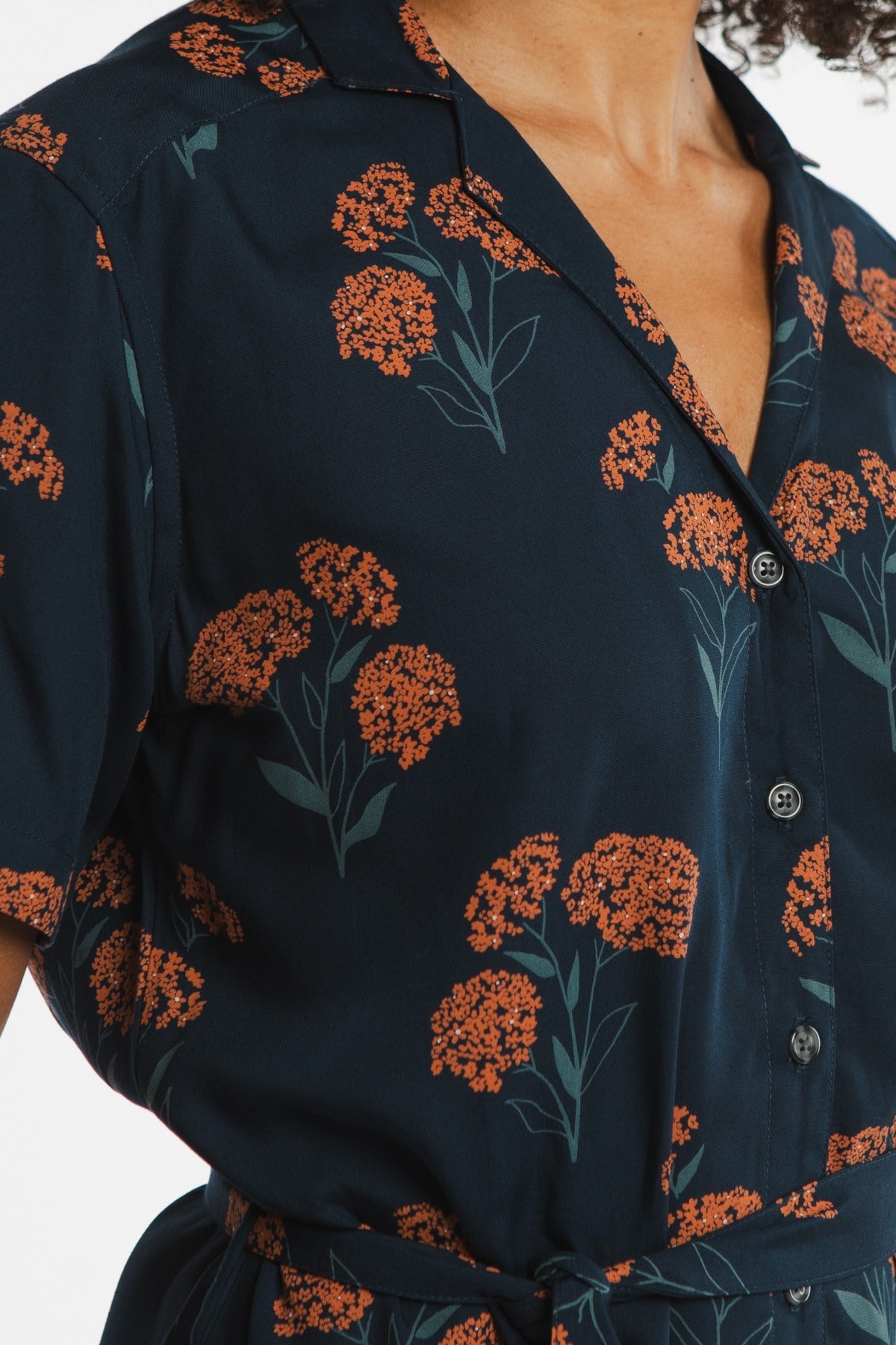 A close-up of a person wearing the Bridge & Burn Sofia Shirt Dress in Wildflower—a navy blue, self-belted, button-front design with orange florals and short sleeves for a stylish, effortless summer look.
