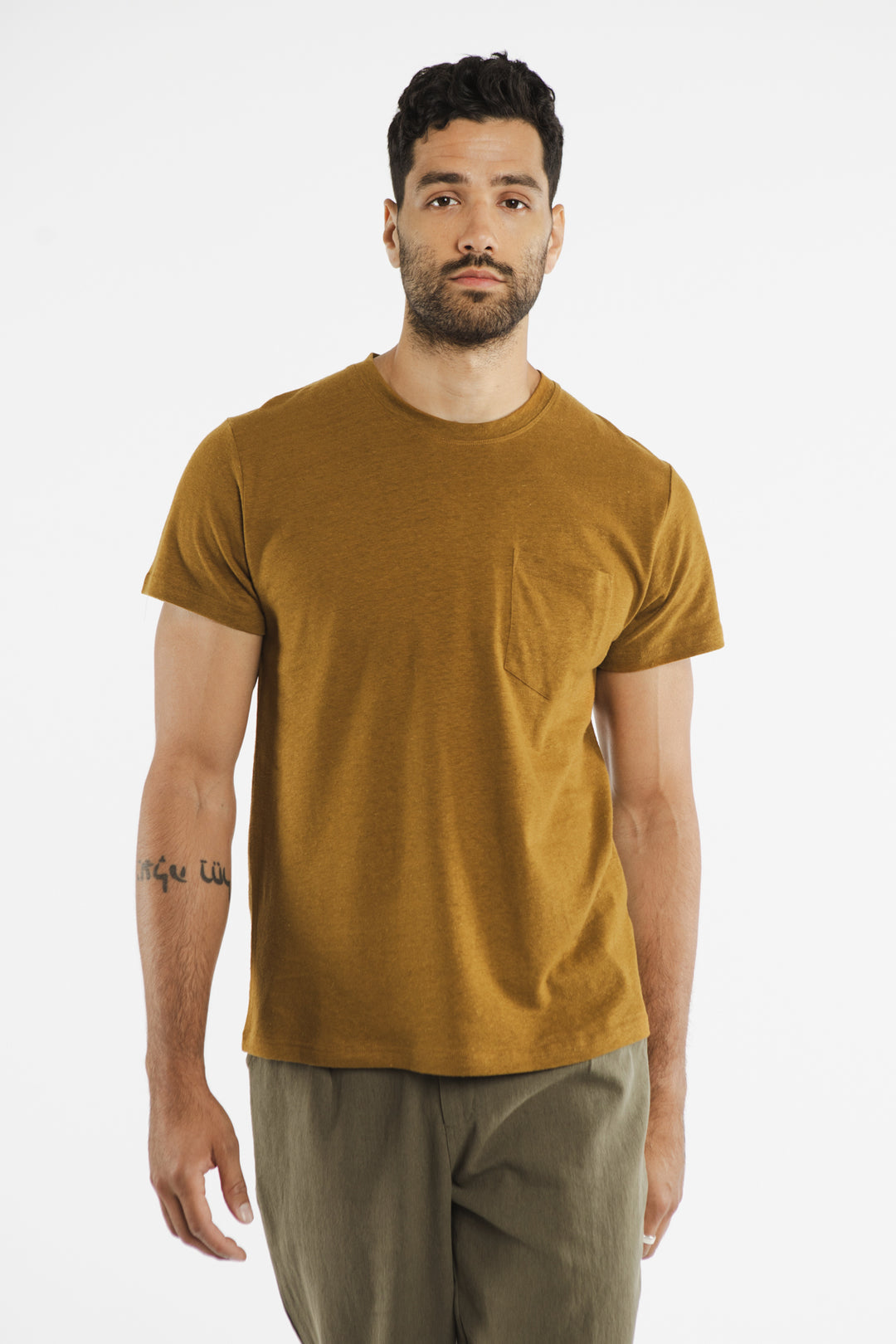 A man with short dark hair and a beard wears the Bridge & Burn Organic Hemp Pocket Tee in Ochre with olive green pants. He stands against a white background, showing a tattoo on his left forearm and a neutral expression.