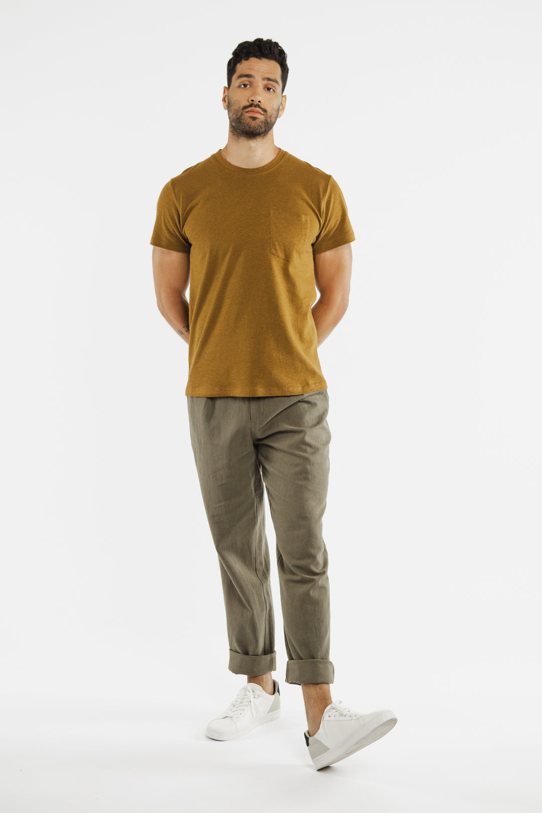 A man with short curly hair stands against a white background, wearing Bridge & Burn’s Organic Hemp Pocket Tee in Ochre, olive green rolled-up pants, and white sneakers, with his hands behind his back.