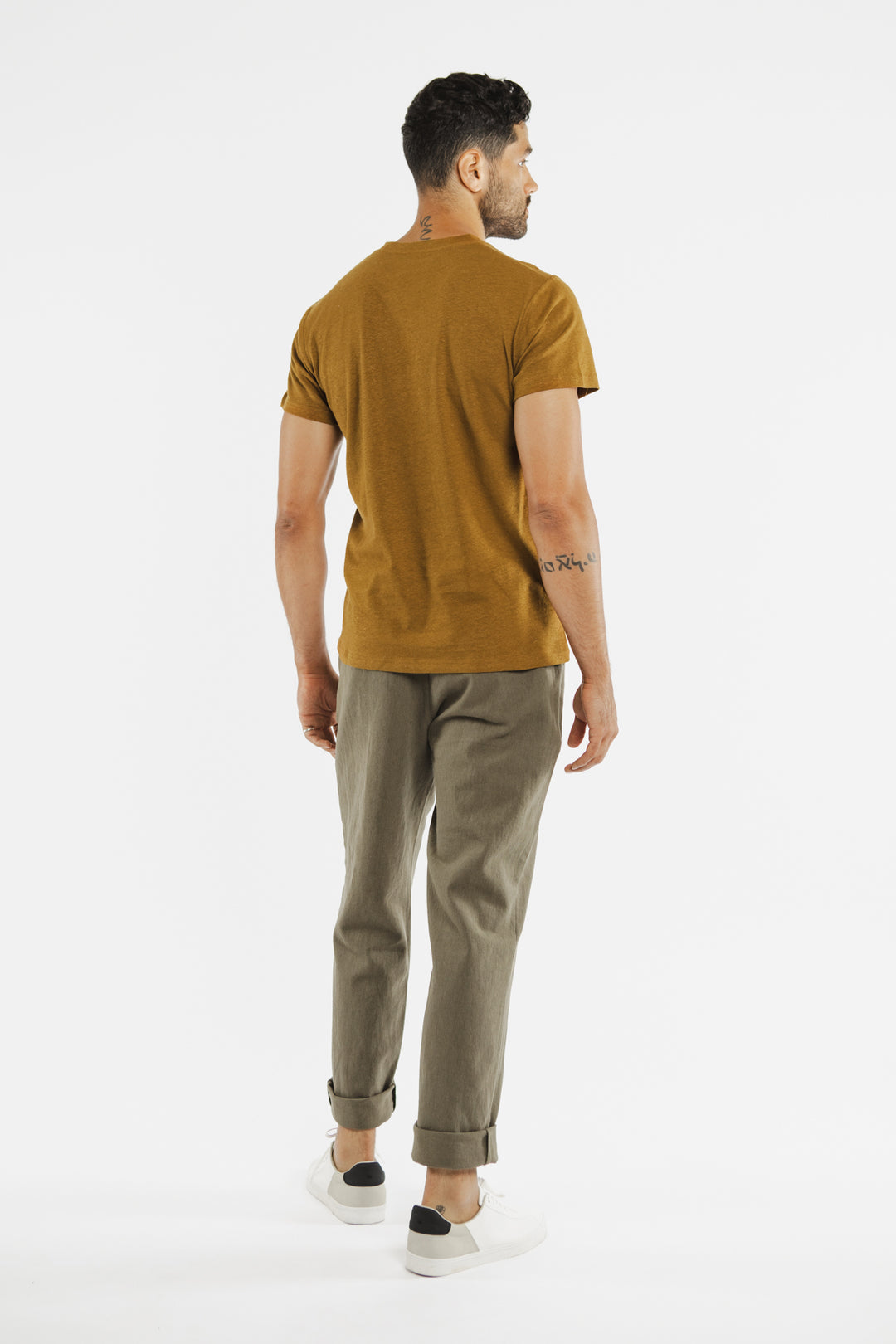 A man stands facing away, wearing the Bridge & Burn Organic Hemp Pocket Tee in Ochre, olive green rolled-up pants, and white sneakers against a plain white background.