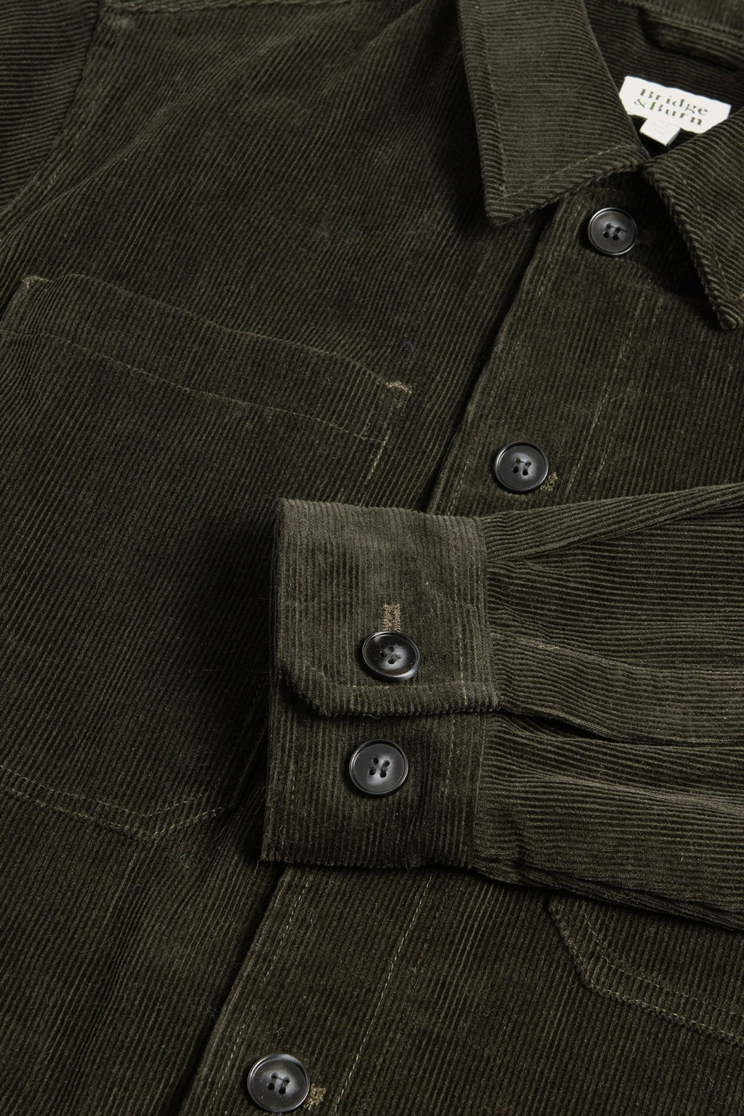 A close-up of the Bridge & Burn Banks Shirt Jacket in Deep Pine Corduroy shows its black buttons, chest pocket, and ribbed texture. One sleeve is folded across the body for a workwear look. The garment label is partially visible at the collar.
