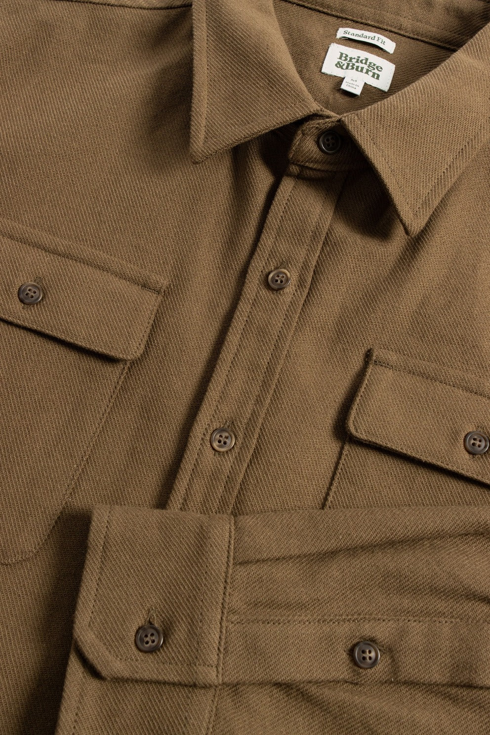 Close-up of the Bridge & Burn Bedford Shirt in olive twill, featuring two chest pockets, brown buttons, and a visible collar tag. The shirt is neatly arranged, showcasing its soft, textured fabric.