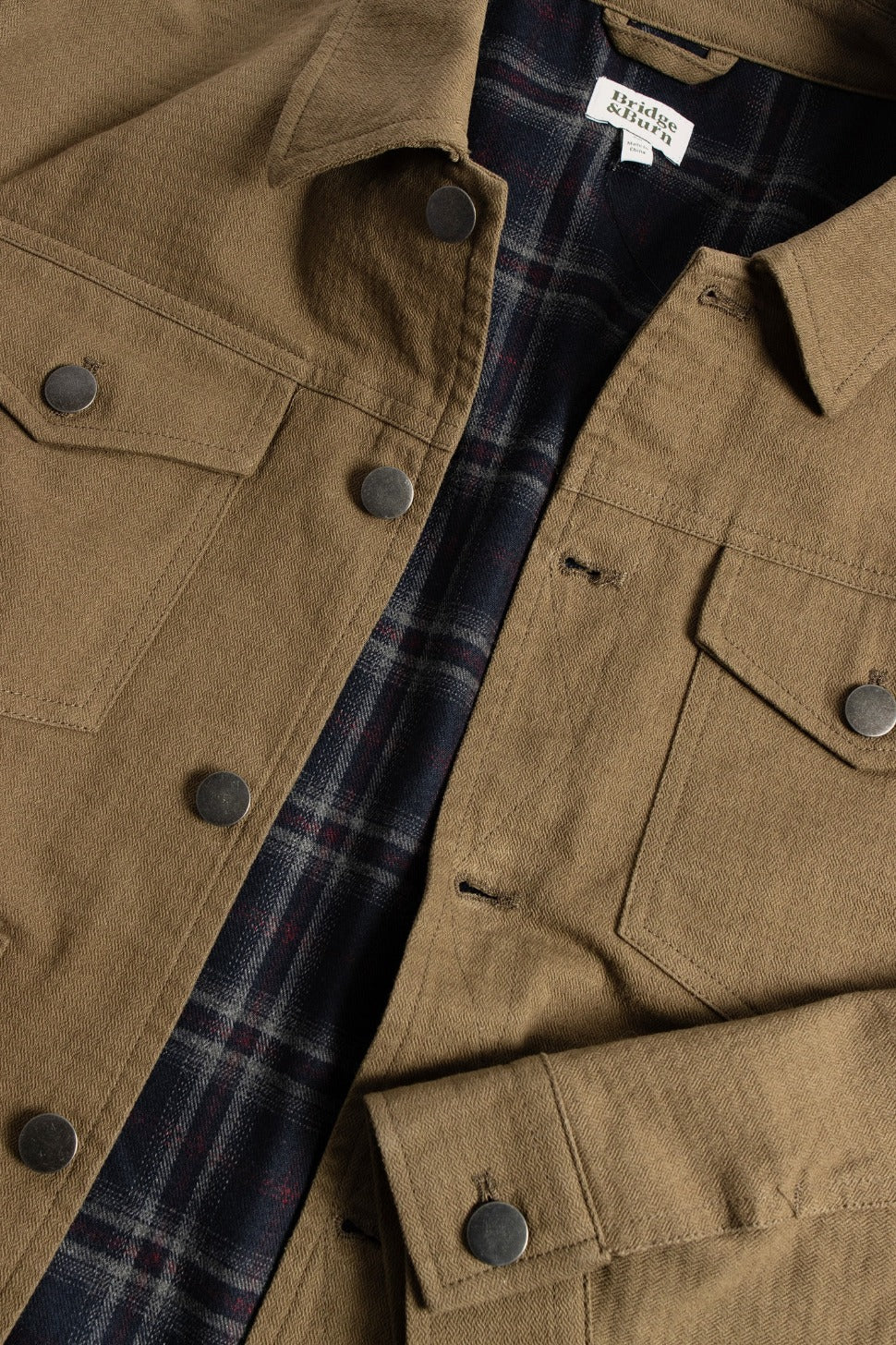 A close-up of the Bridge & Burn Burl Trucker Jacket in Warm Olive, featuring black snap buttons, flap chest pockets, and an inner flannel lining with a dark blue and red plaid pattern.