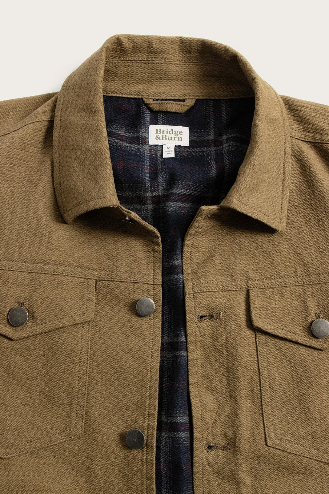 The Bridge & Burn Burl Trucker Jacket in warm olive features flap chest pockets, metal buttons, and a plaid flannel lining.