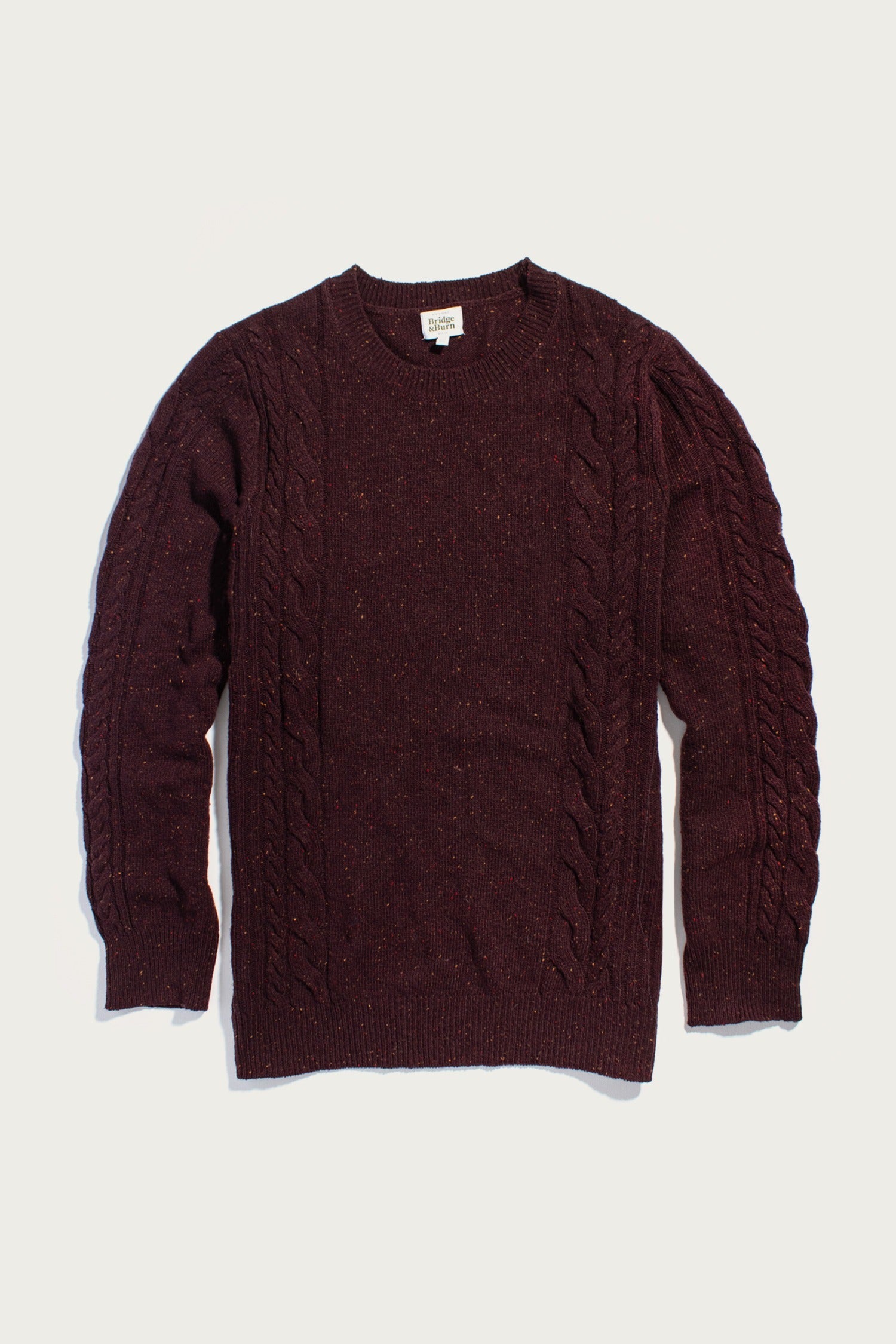 The Hugo Sweater by Bridge & Burn in Burgundy Donegal is a maroon cable-knit crewneck with long sleeves, ribbed cuffs and hem, crafted from sustainable wool, featuring visible cable detail on the front and sleeves.