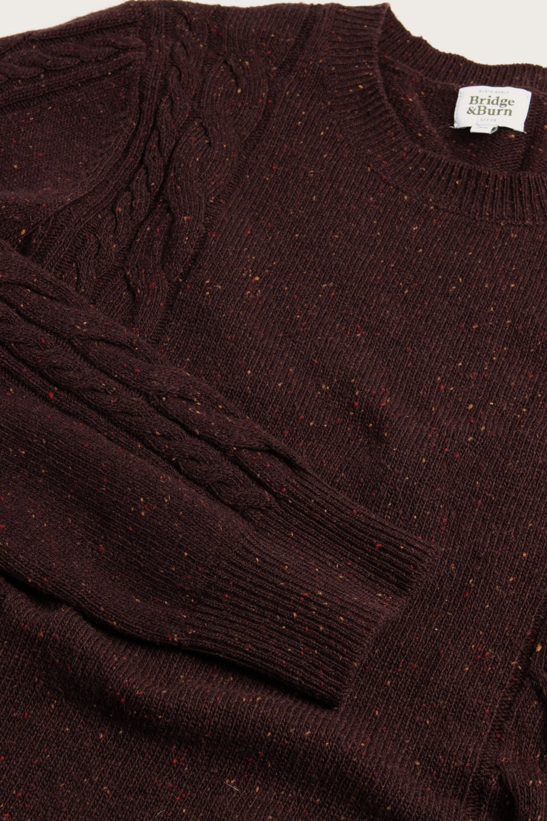A close-up of the Bridge & Burn Hugo Sweater in Burgundy Donegal—a crewneck with dark speckles, cable-detailed sleeves, sustainable wool, and a visible brand label at the neck.