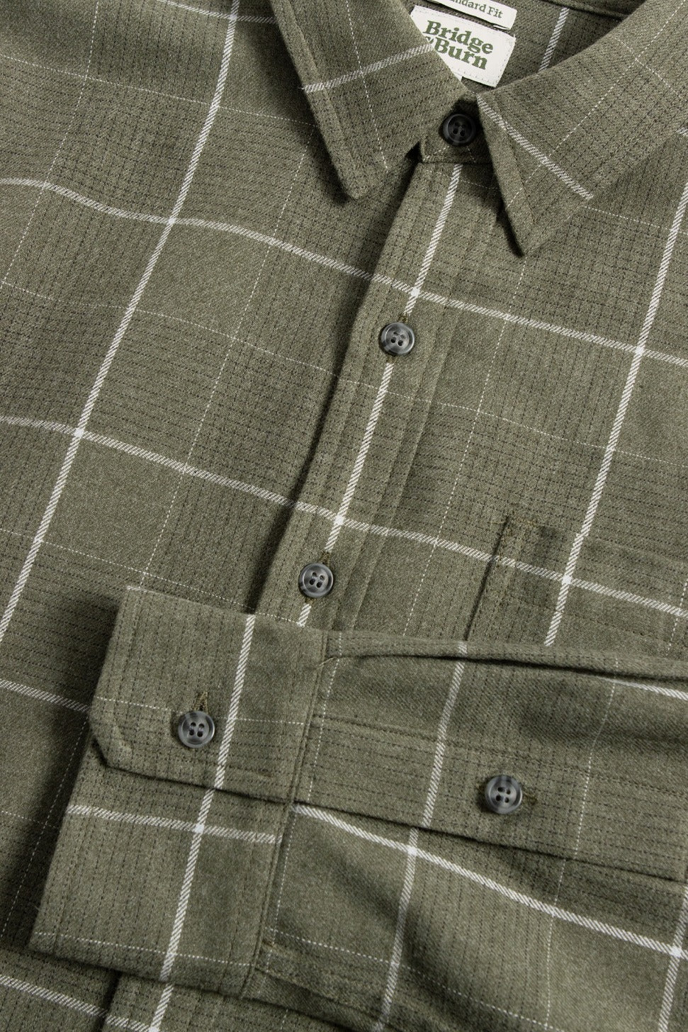 Close-up of the Larson Shirt / Rosemary Twill Windowpane by Bridge & Burn, an olive green plaid button-up with white lines, front pocket, buttoned cuffs, dark grey buttons, and a Bridge & Burn label inside the collar.