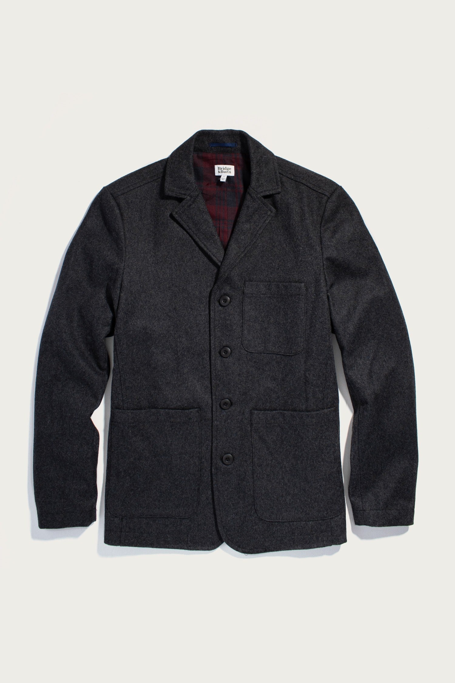 The Nate Chore Blazer in Onyx Wool by Bridge & Burn is a dark charcoal gray wool-blend blazer with a notched collar, button front, and three pockets. Laid flat on white, its dark red plaid lining adds a stylish winter touch.