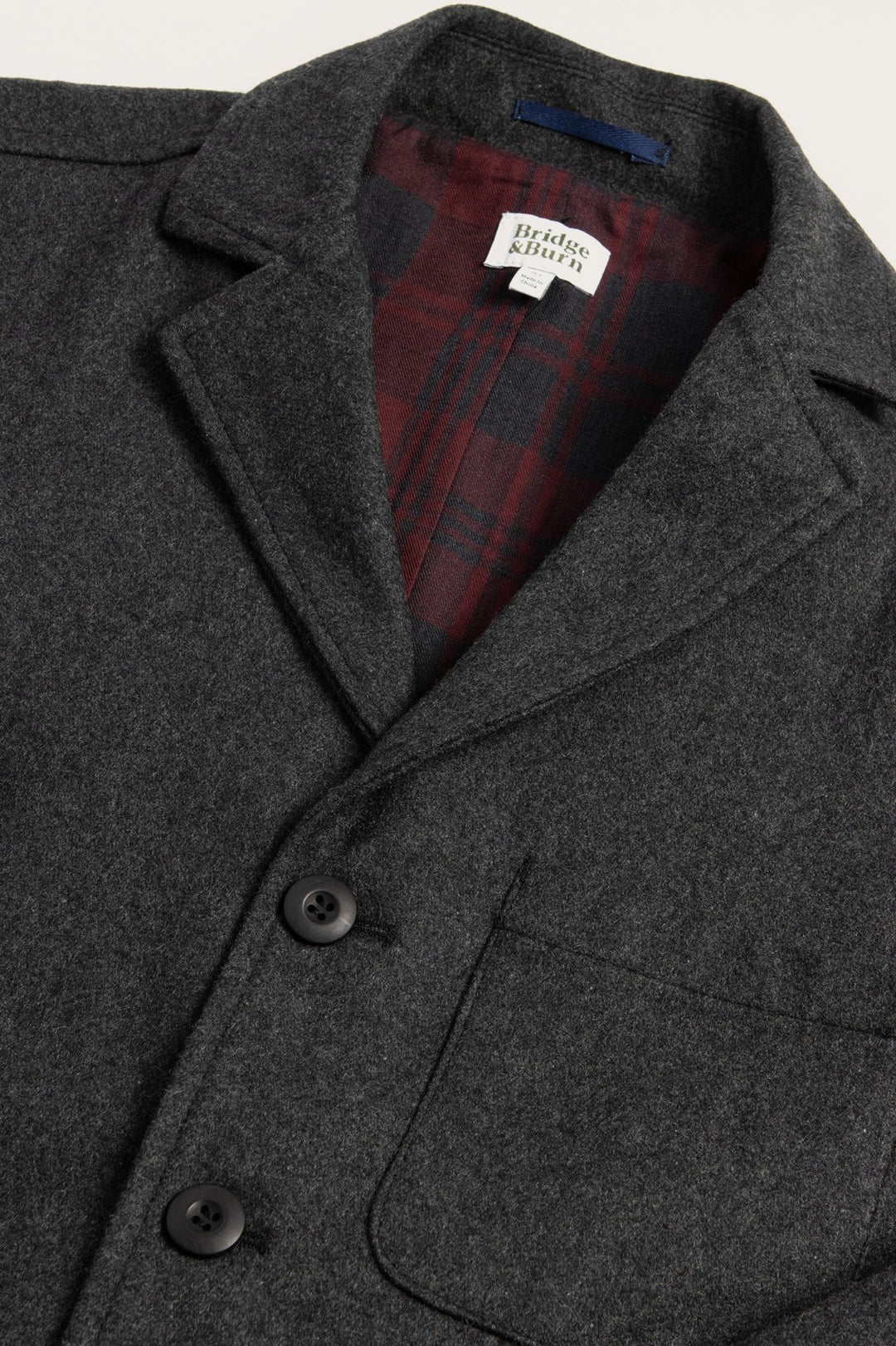 A close-up of the Bridge & Burn Nate Chore Blazer in Onyx Wool shows dark grey wool-blend fabric, black buttons, a front pocket, and visible red-and-black plaid lining with a white label stitched near the collar.