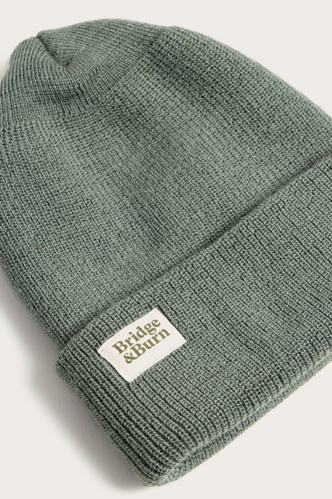 The Bridge & Burn Wool Watch Cap / Foliage is a sage green knit cap with a folded cuff, made in the USA from 100% wool, and features a white rectangular Bridge & Burn label sewn on the front.