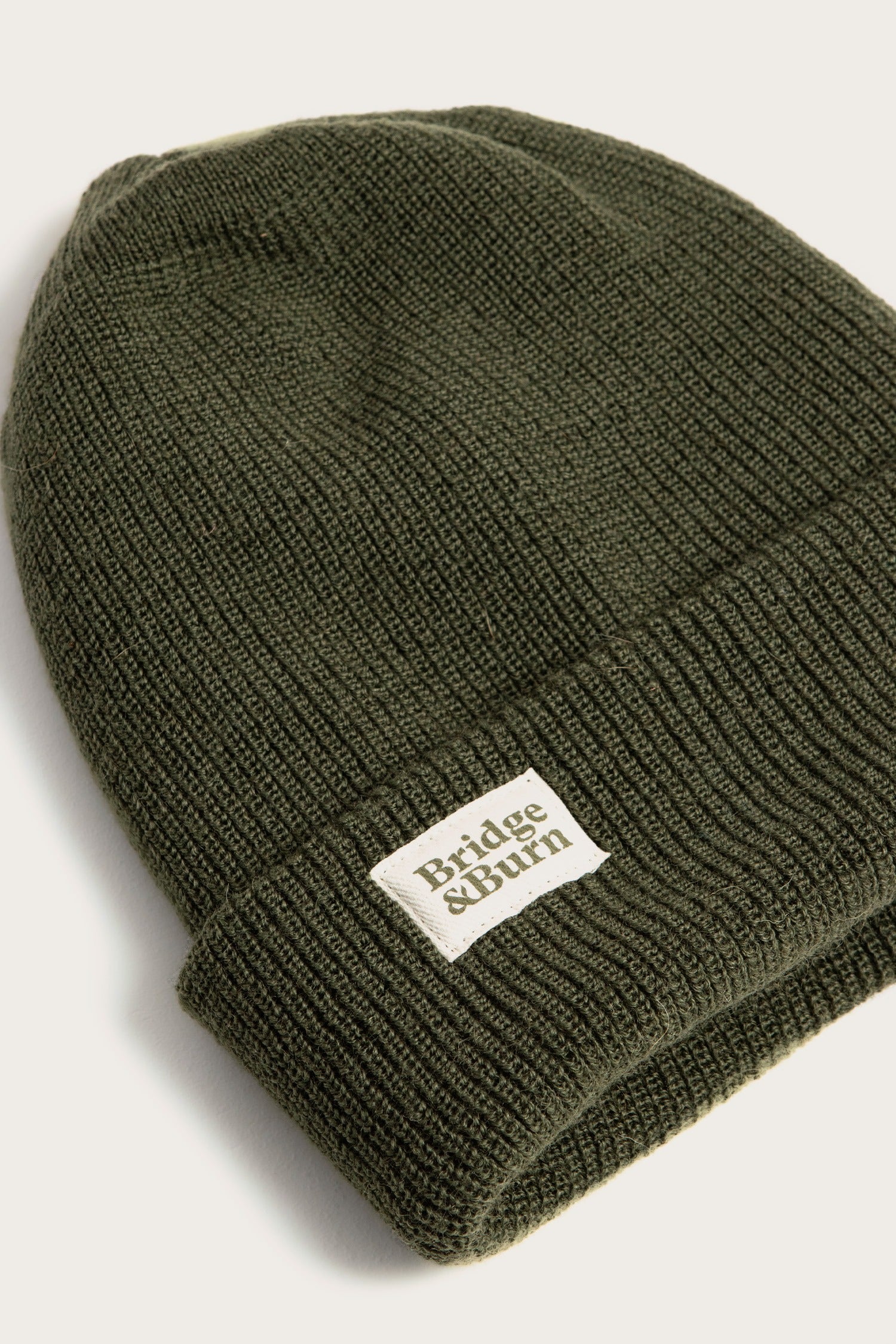 The Bridge & Burn Wool Watch Cap in Olive Drab is 100% wool, made in the USA, and features a folded brim with a white rectangular brand patch on the front. Shown on a plain light background.