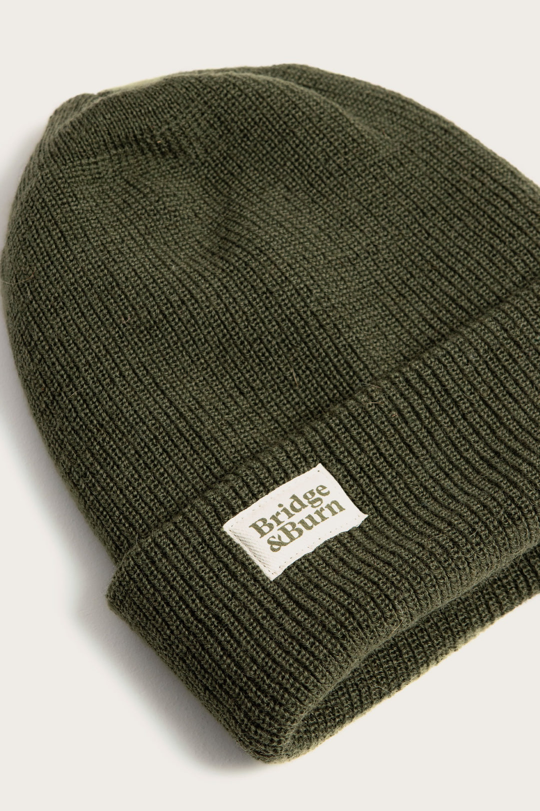 The Bridge & Burn Wool Watch Cap in Olive Drab is 100% wool, made in the USA, and features a folded brim with a white rectangular brand patch on the front. Shown on a plain light background.