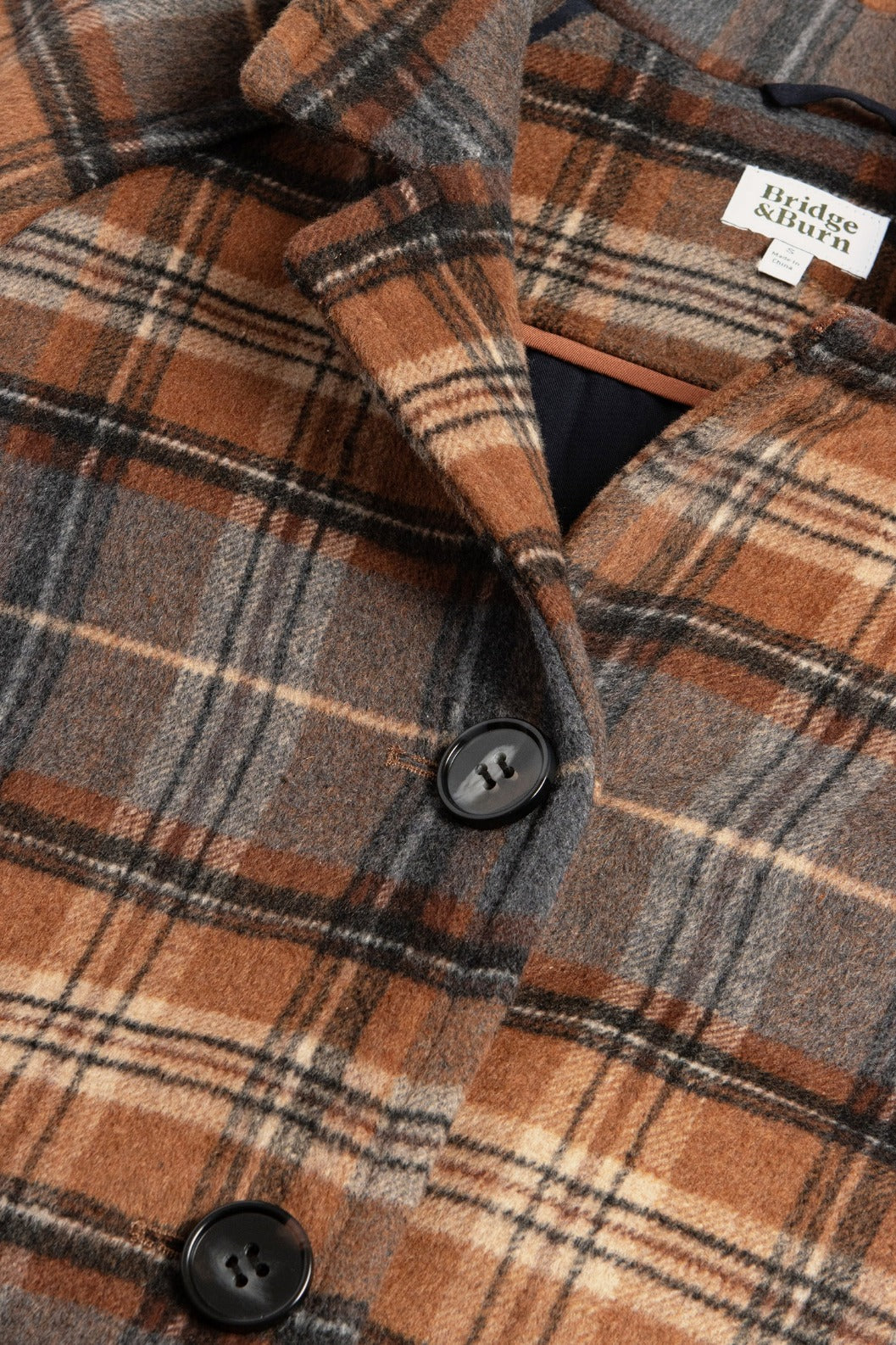 Close-up of the Alberta Car Coat in Dorset Brushed Plaid by Bridge & Burn, featuring a brown, gray, and cream plaid wool blend, large black buttons, and a visible brand label near the collar.