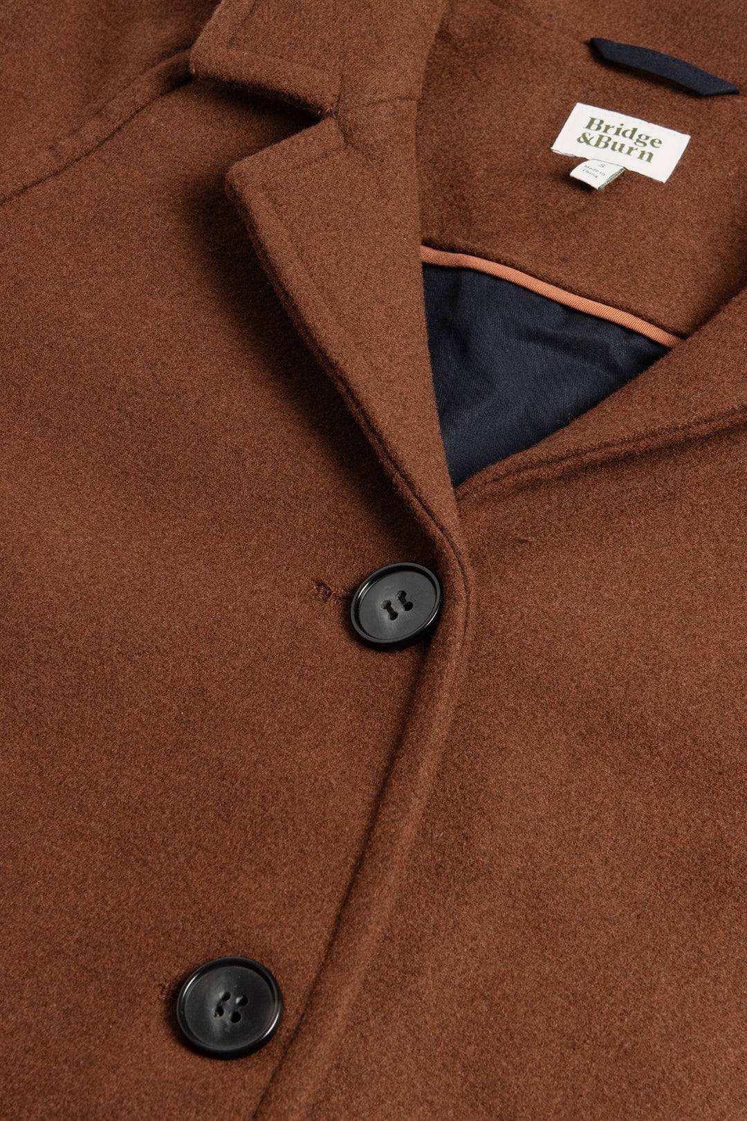 Close-up of the Alberta Car Coat in Fired Brick by Bridge & Burn, featuring black buttons, a notched collar, and visible label. The partially open coat reveals its dark brushed wool blend lining with tan accent trim.