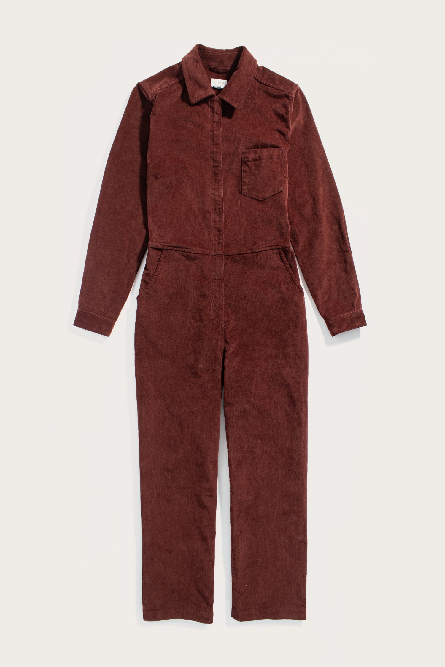 The Bridge & Burn Faye Jumpsuit in Dark Red Pinwale, a women’s corduroy style with long sleeves, collar, chest and side pockets, and workwear-inspired details, is laid flat on a plain white background.