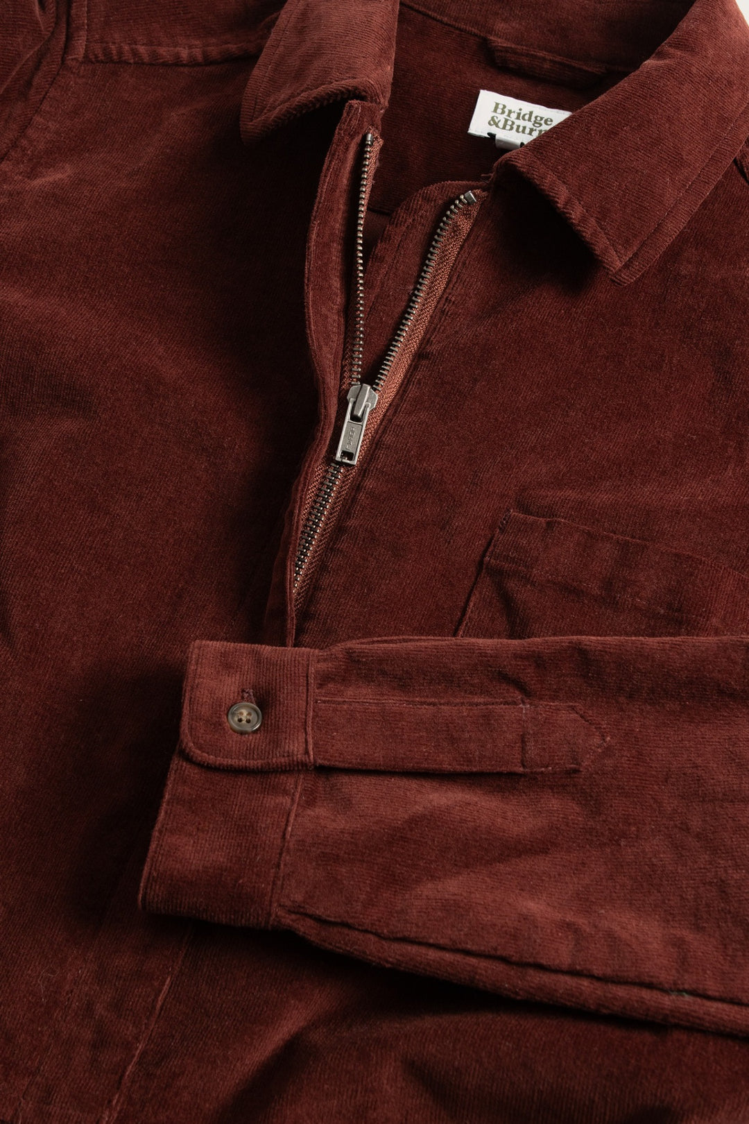 A close-up of the Bridge & Burn Faye Jumpsuit in Dark Red Pinwale highlights its corduroy fabric, chest pocket, zip front, and buttoned cuffs for a classic workwear-inspired look with detailed stitching.