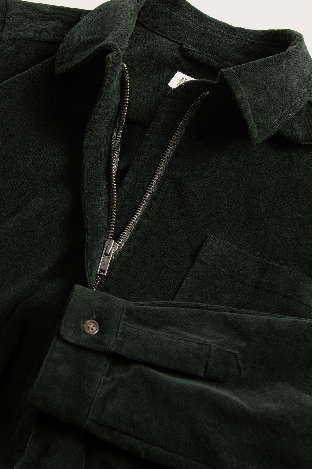 A close-up of the Bridge & Burn Faye Jumpsuit in Deep Pine Pinwale, a dark green stretch corduroy style with a half-zip front, chest pocket, and buttoned sleeve cuffs.