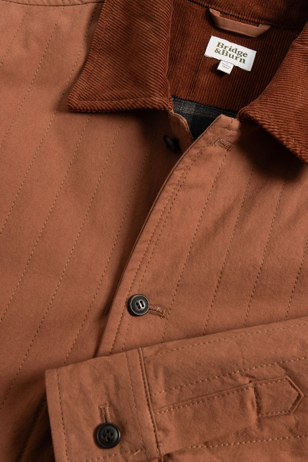 Close-up of the Kenton Jacket in Sienna—a brown quilted jacket with a corduroy collar, black buttons, and a white label inside the collar showing the Bridge & Burn brand. This lightweight piece offers both style and comfort.