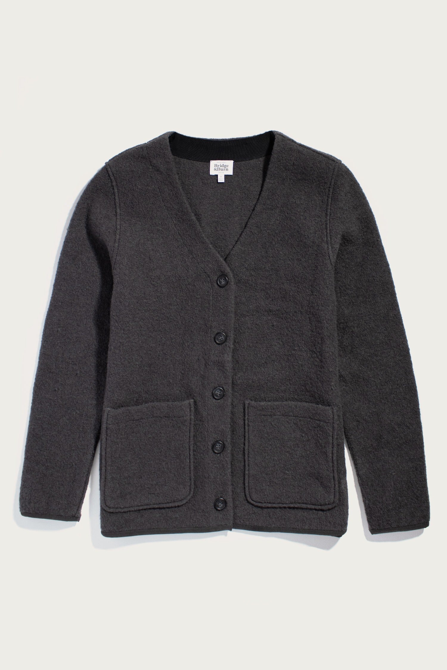 The Bridge & Burn Whit Cardigan in Charcoal is a dark gray, long-sleeve boiled wool cardigan with a V-neck, five black front buttons, and two large patch pockets, shown laid flat on a light background.