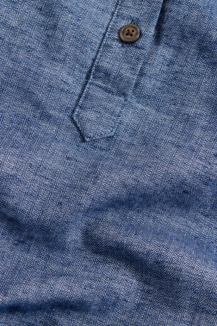 Close-up of the Yarra Pullover by Bridge & Burn in indigo herringbone denim, showing a chevron weave pattern with part of the shirt placket and a single brown button.